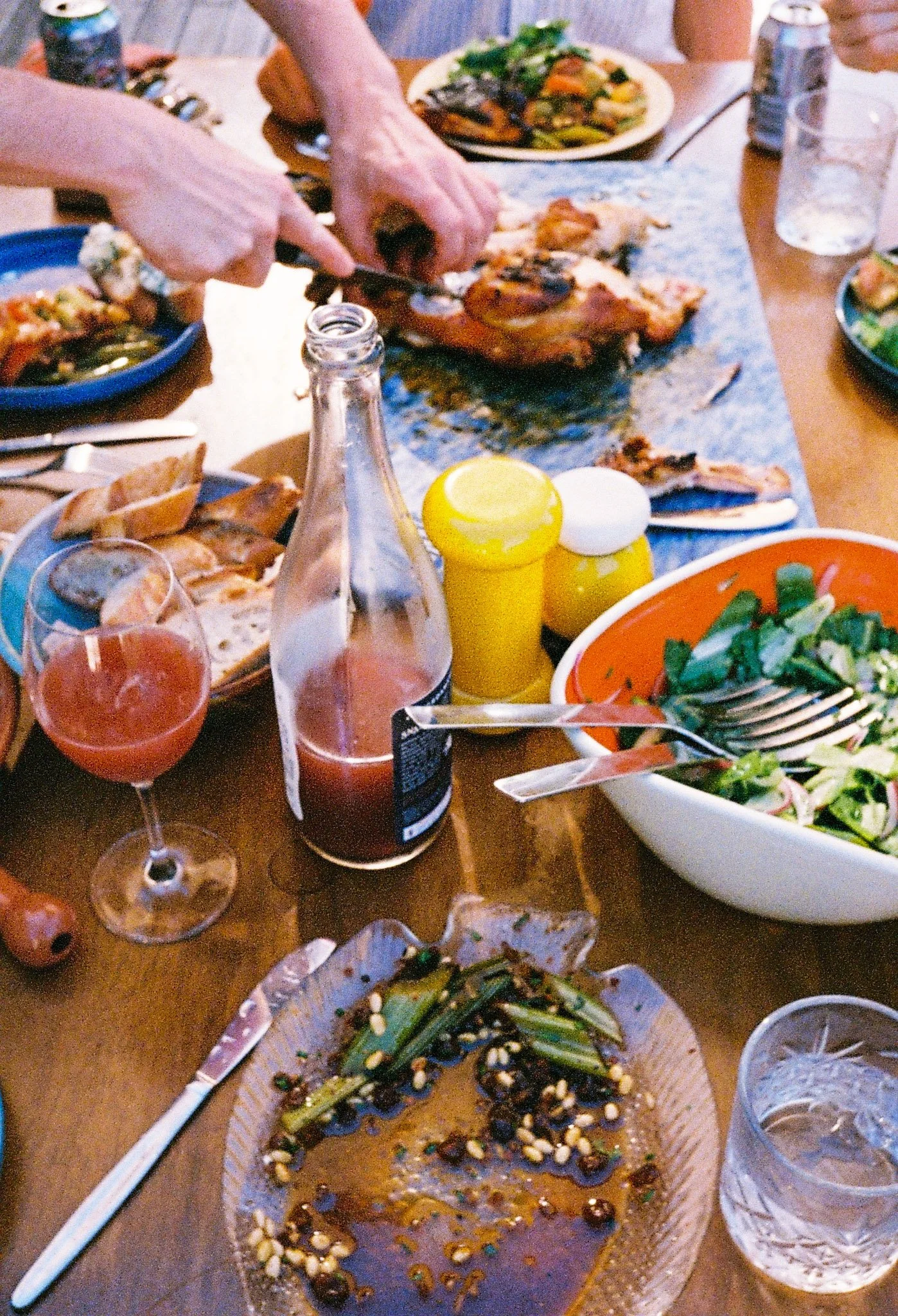 A table with food, a partially eaten salad, a bottle of wine, condiments, and person carving barbecue chicken off a blue chopping board.