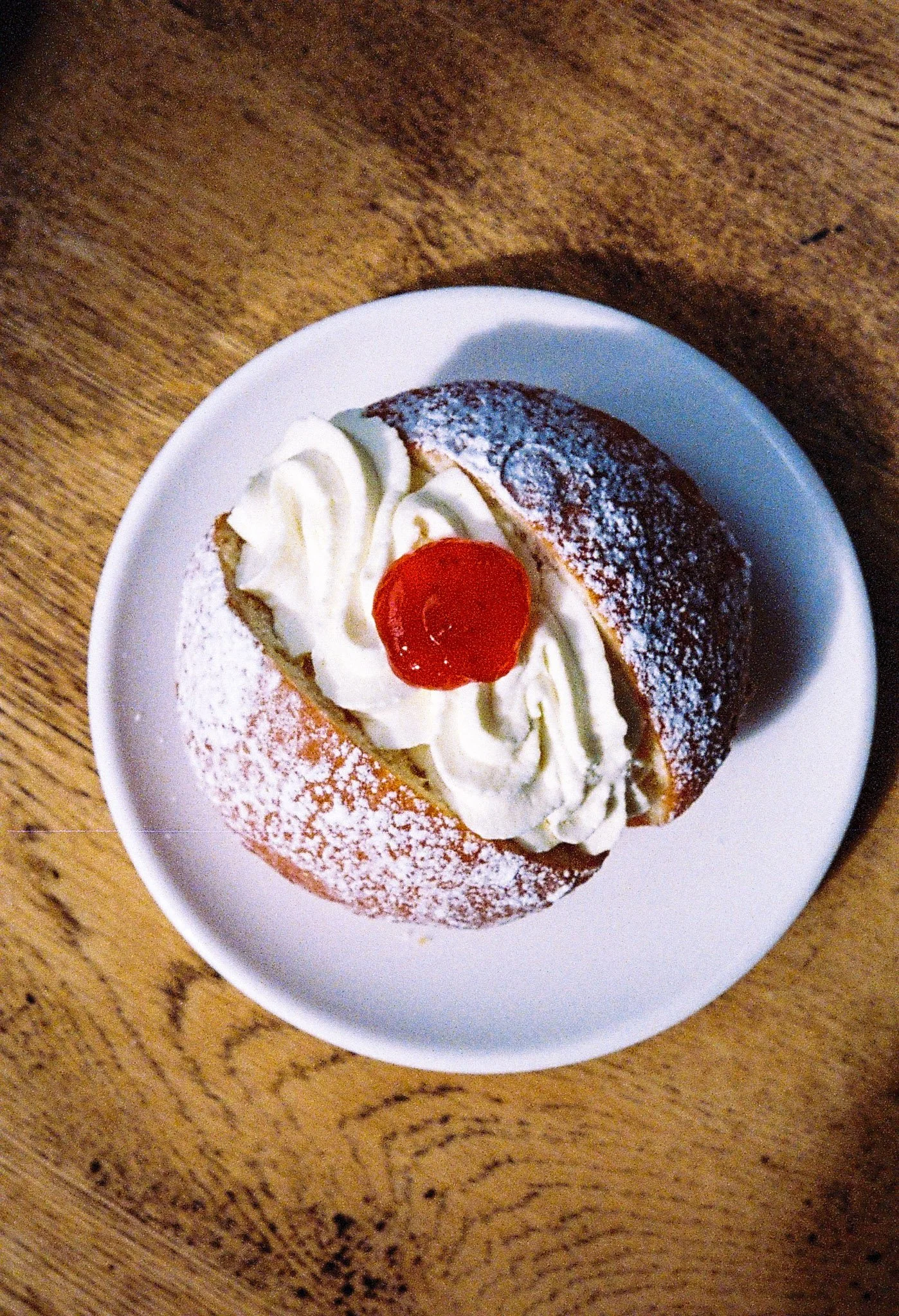 A cream-filled pastry with powdered sugar, whipped cream, and a cherry on top, served on a white plate on a wooden table.