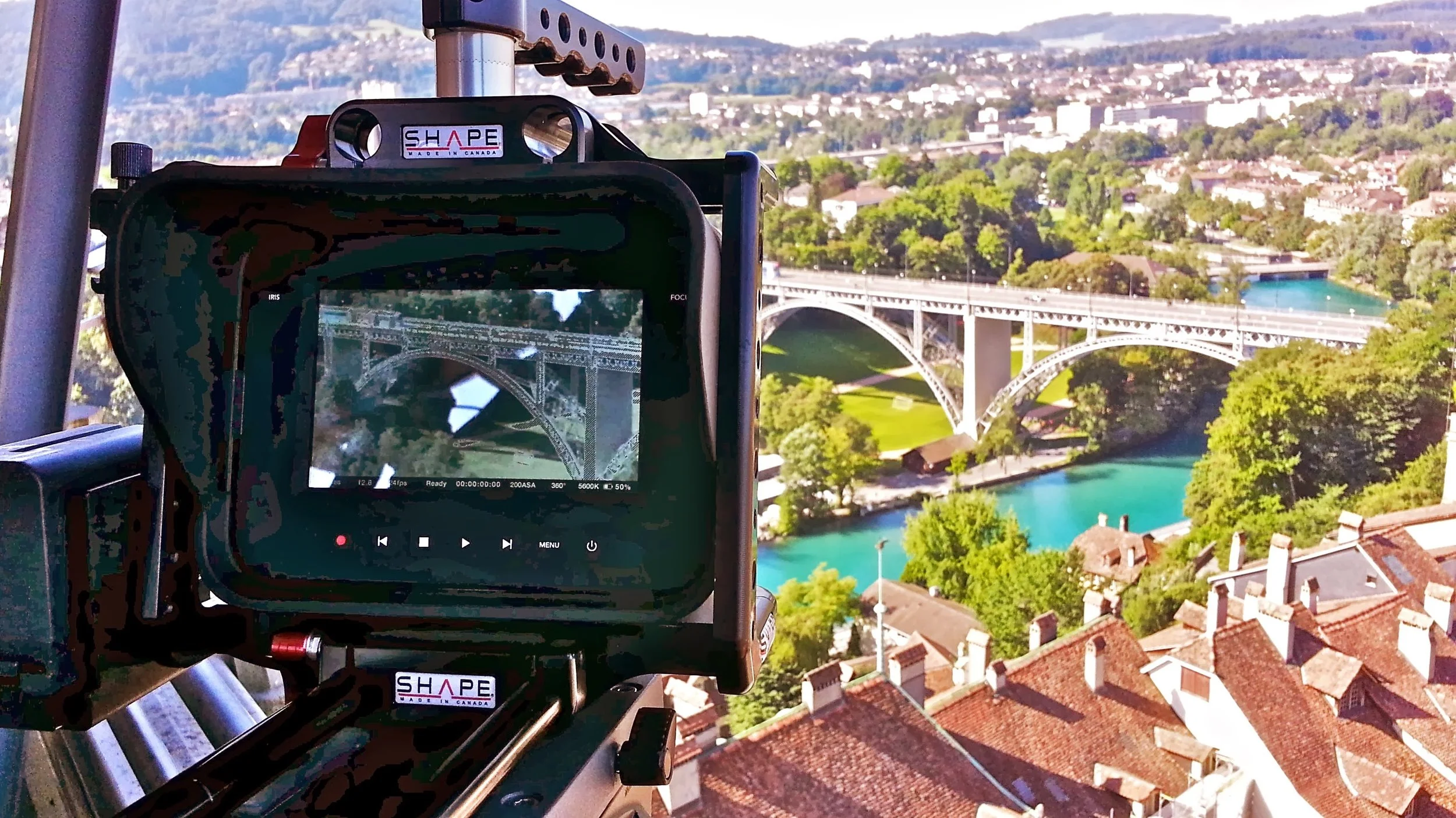 The image shows a camera mounted on a tripod, capturing a view of a steel arch bridge over a river, with a cityscape and greenery in the background. The camera's screen displays a live preview of the bridge.