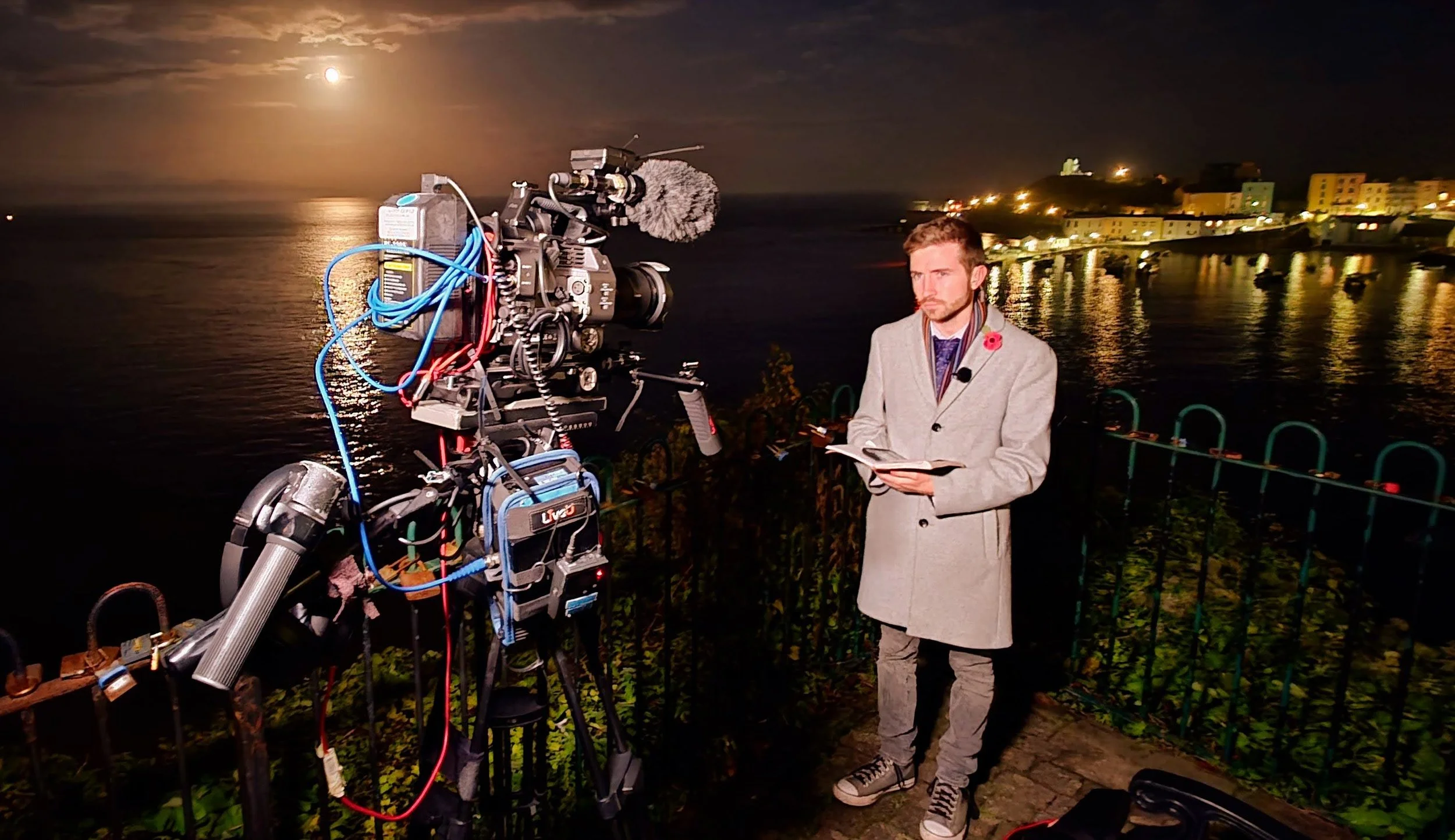 A man in a gray coat holding a notebook stands near a large film camera by a body of water at night with the moon visible in the sky and illuminated buildings reflecting on the water.