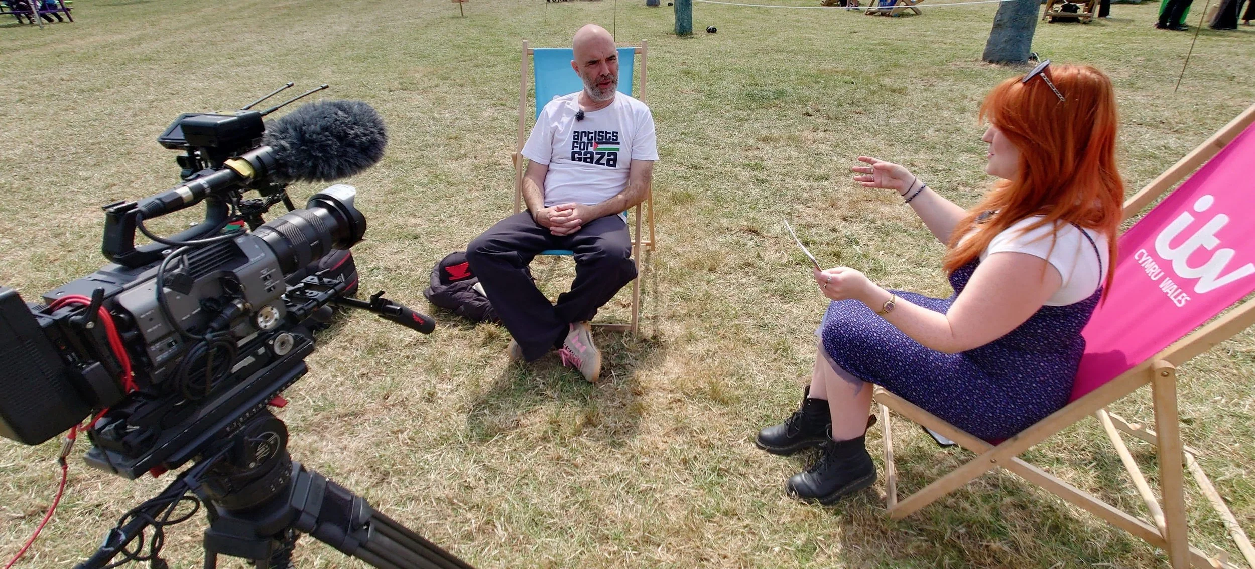 A man being interviewed outdoors in a grassy area, sitting on a folding chair. A woman with red hair, sitting on a pink chair, is interviewing him and holding a card. A professional video camera is filming the interview.