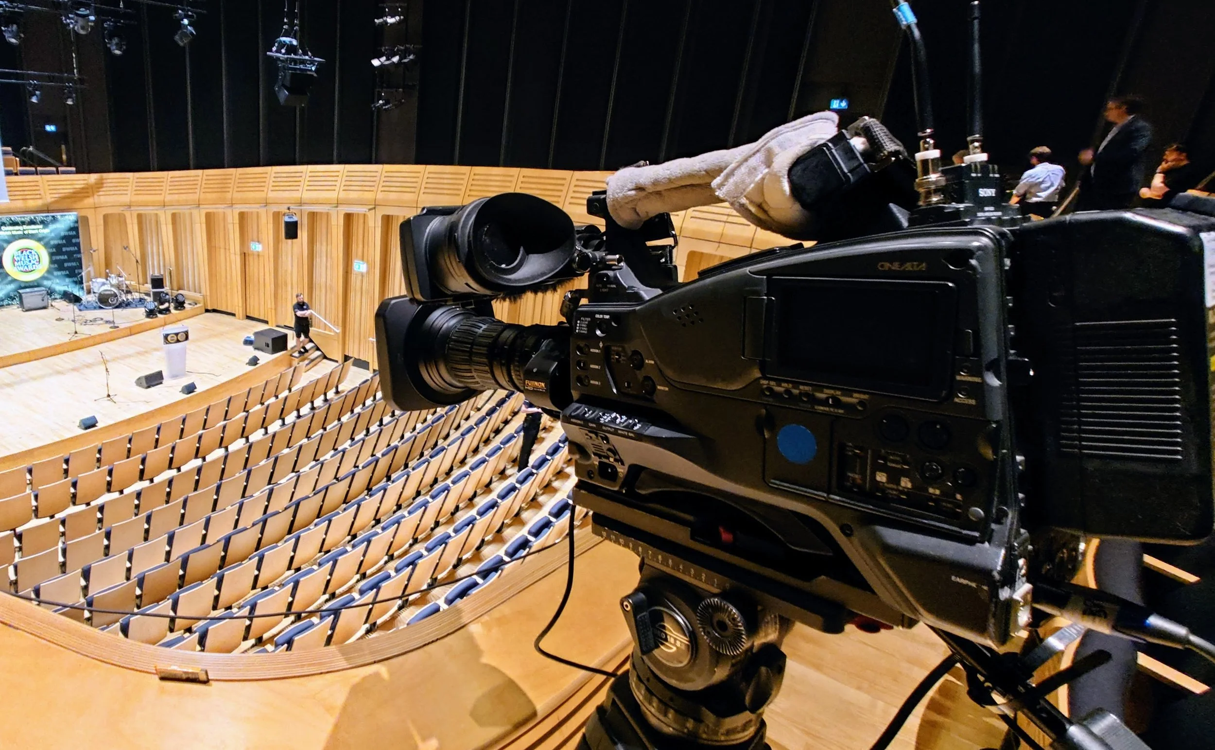 Close-up of a professional video camera on a tripod, set up inside an empty auditorium with wooden seats, a stage with musical instruments, and a few people and crew members preparing for a performance.