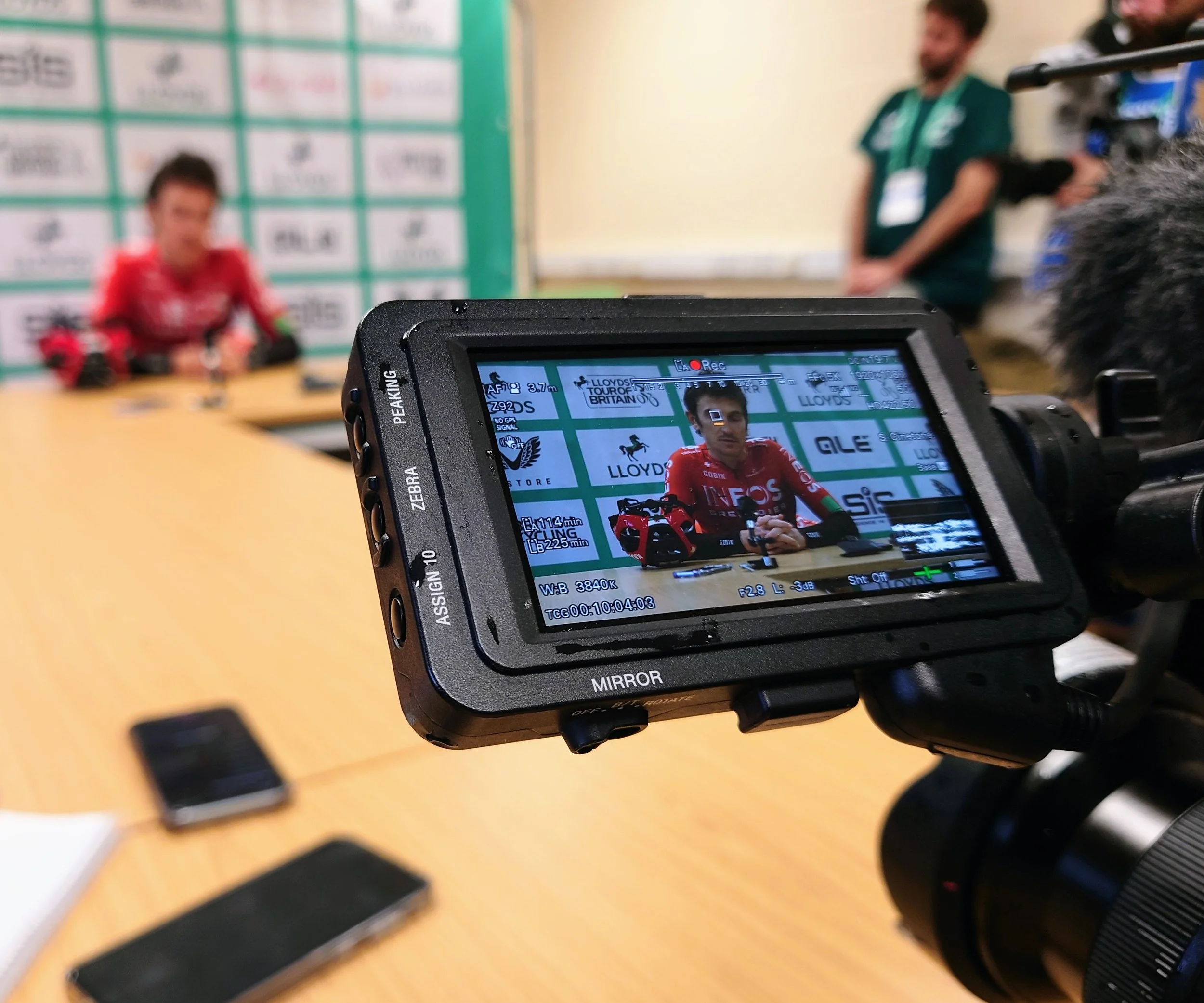Camera view showing a cyclist in a red and black team jersey being interviewed at a press conference, with a blurred background and several smartphones on a table.