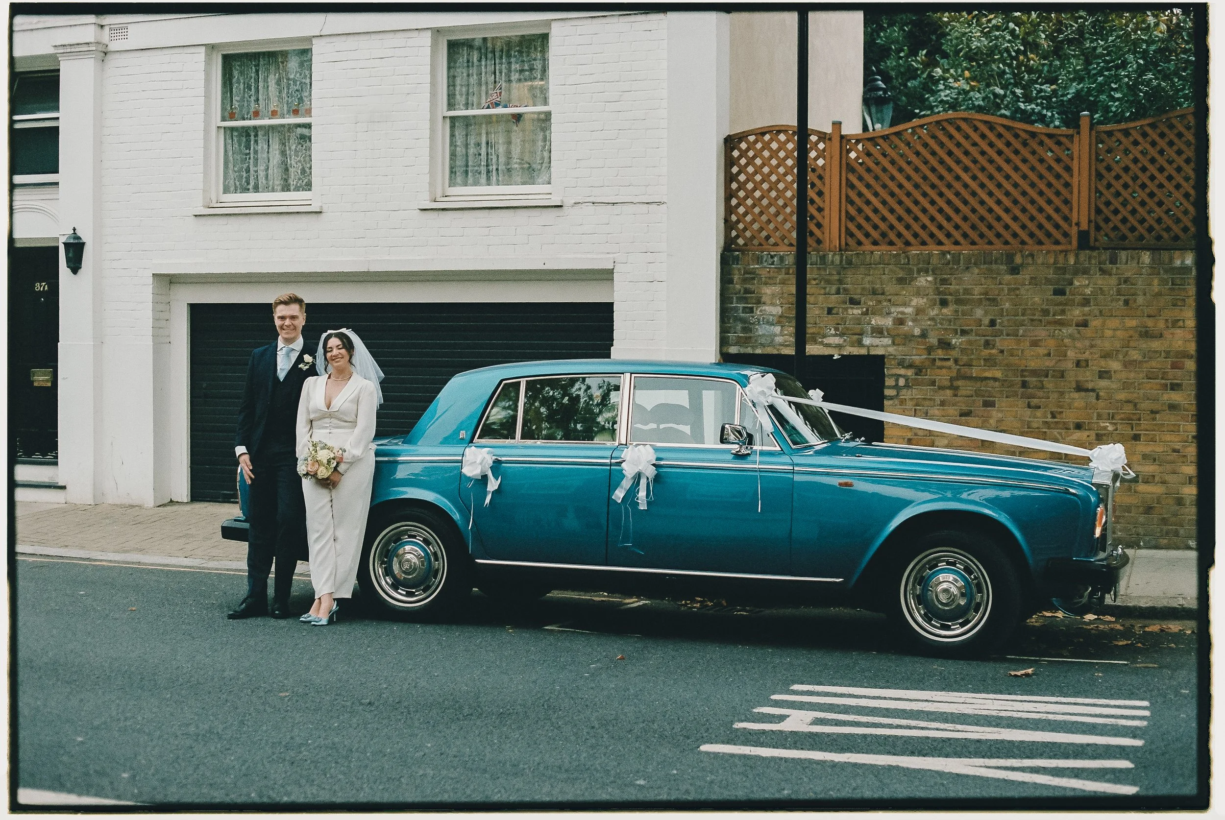 A newlywed couple standing beside a decorated blue vintage Rolls Royce on a city street, in front of a white building with windows and a garage door.