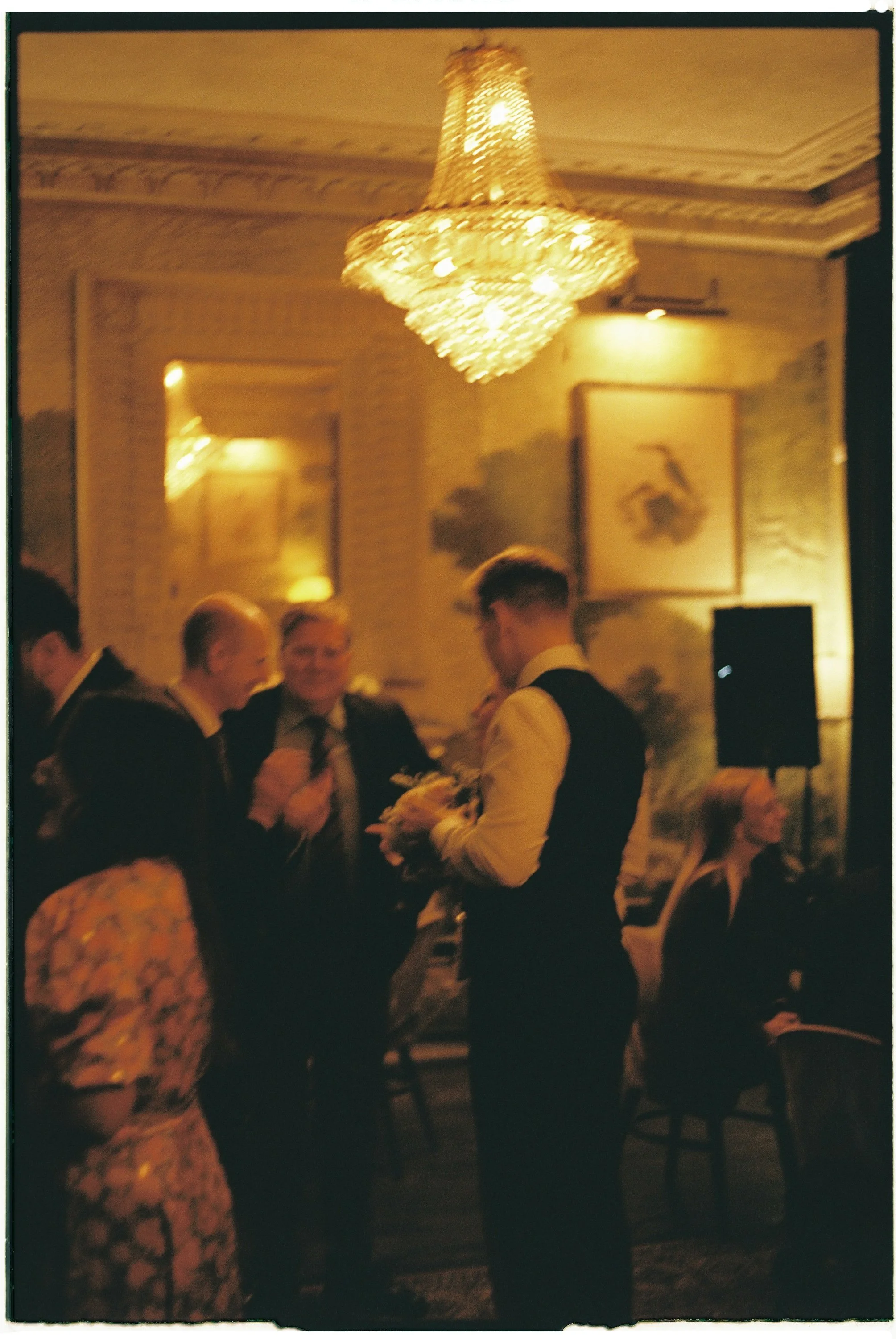 A chandelier hanging from an ornate ceiling illuminates a dimly lit room where a group of people in formal attire are engaged in conversation, with some seated and others standing.