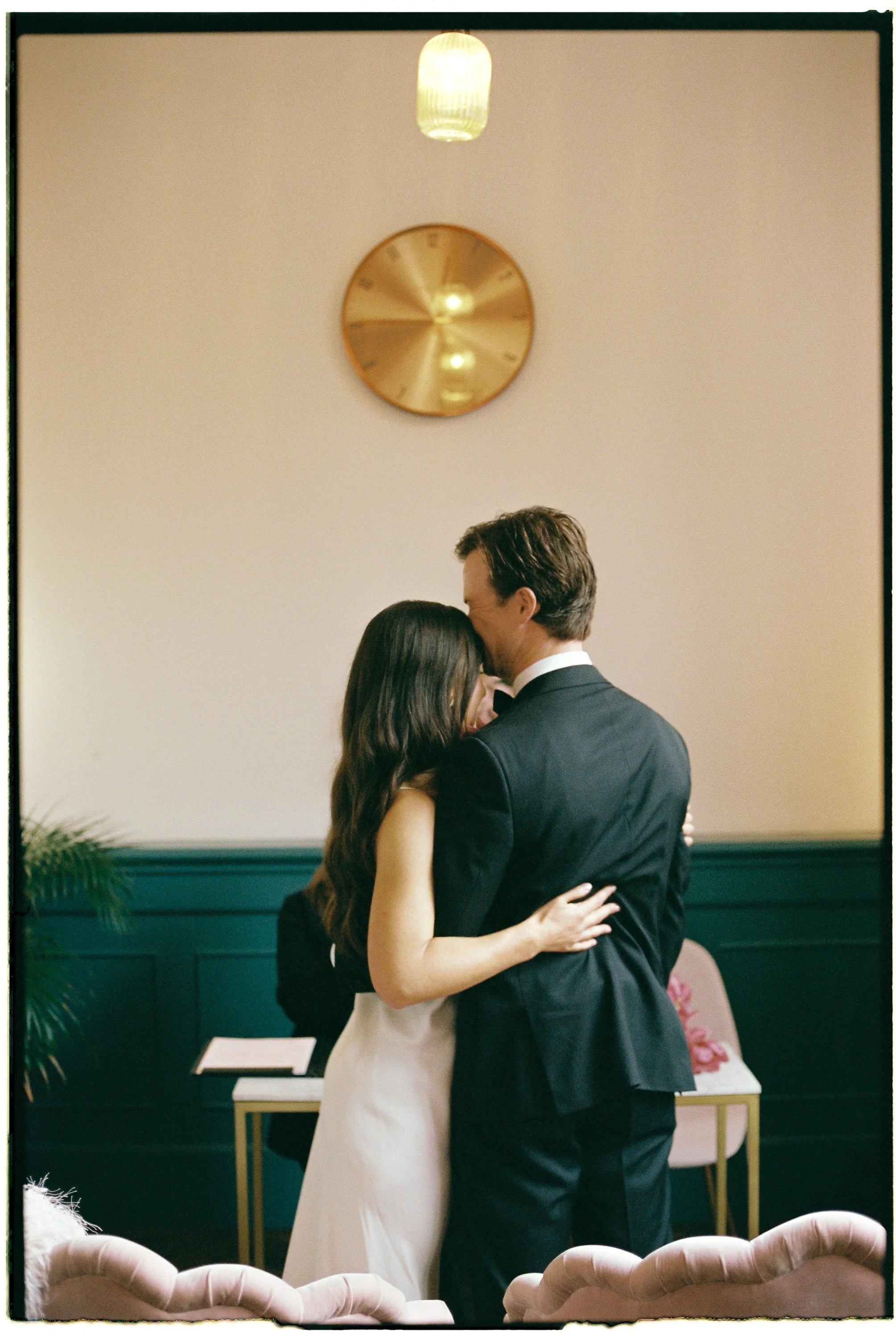 A couple dancing closely at a formal event, with the woman wearing a white dress and the man wearing a black suit, in a room with green walls and a large gold clock on the wall.