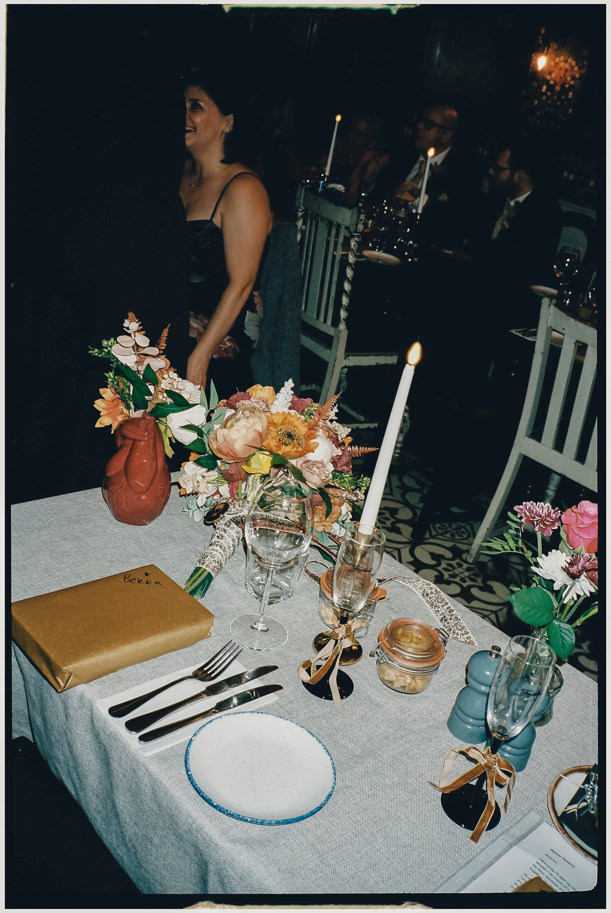 A decorated dining table with floral arrangements, candles, glassware, and wrapped gifts, set for a celebration in a dimly lit venue, with people seated in the background.