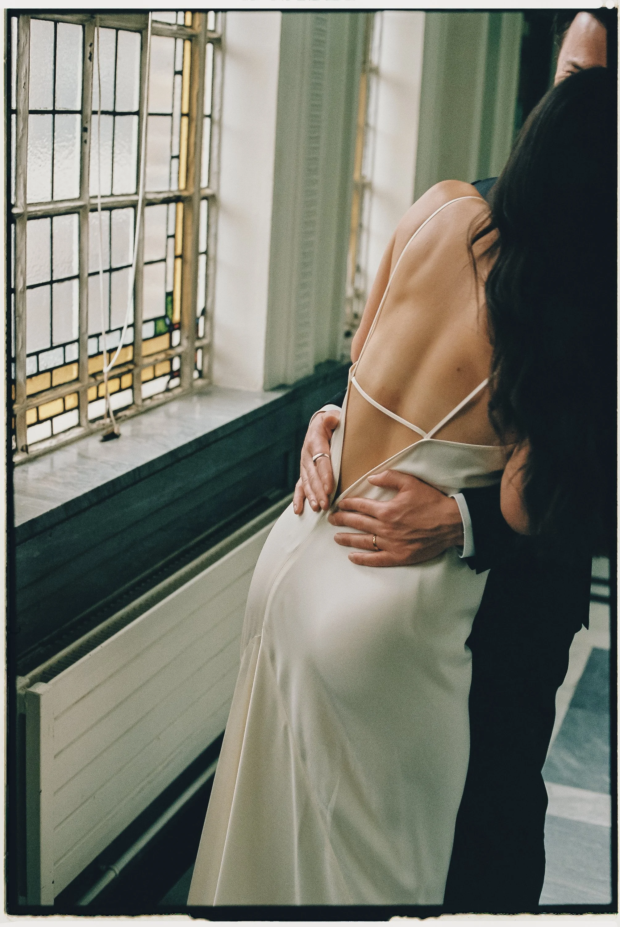 A bride in a backless wedding dress is embraced by a man in a suit, in front of a stained glass window.