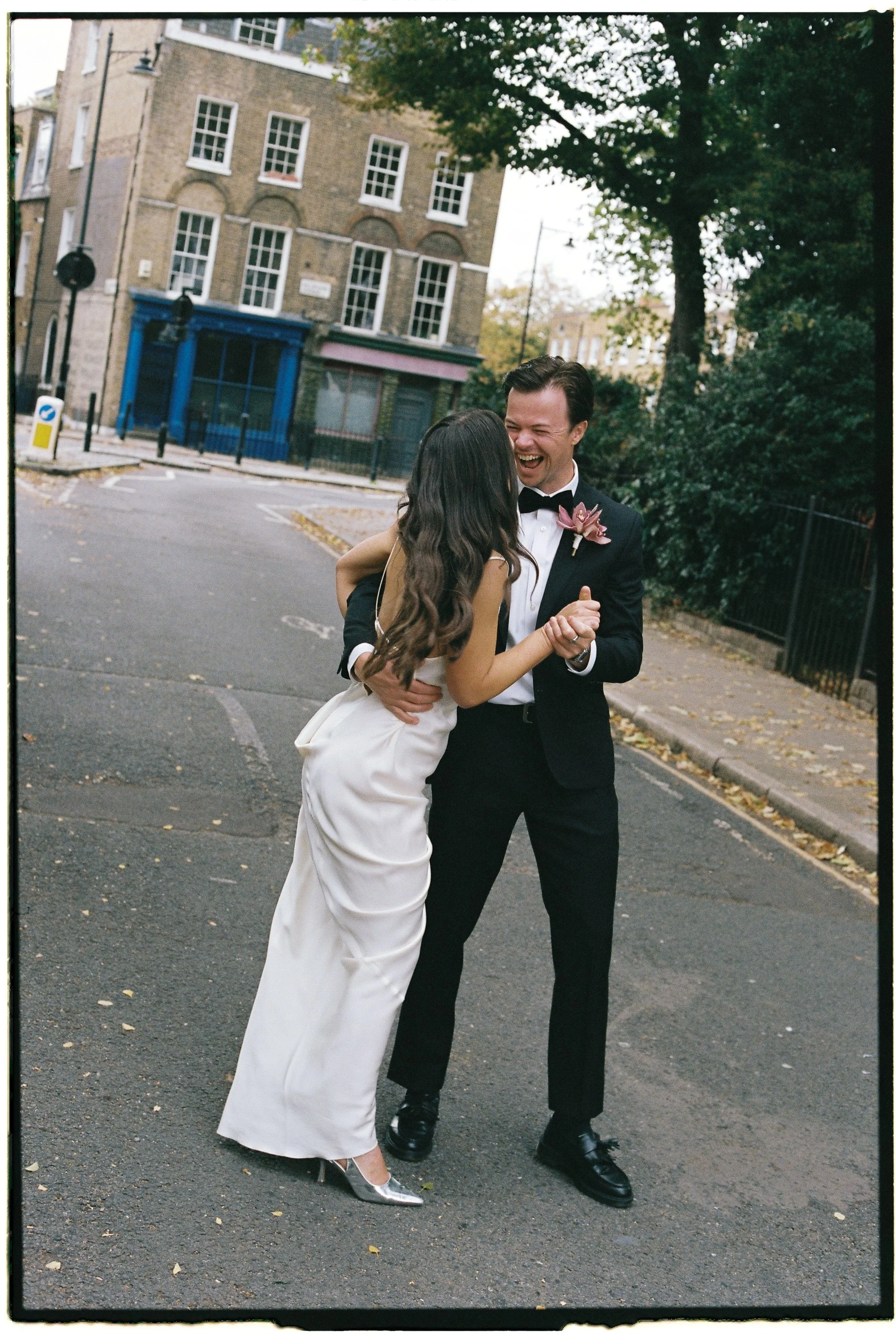 A couple in wedding attire dancing and laughing outdoors on a street with trees and buildings in the background.