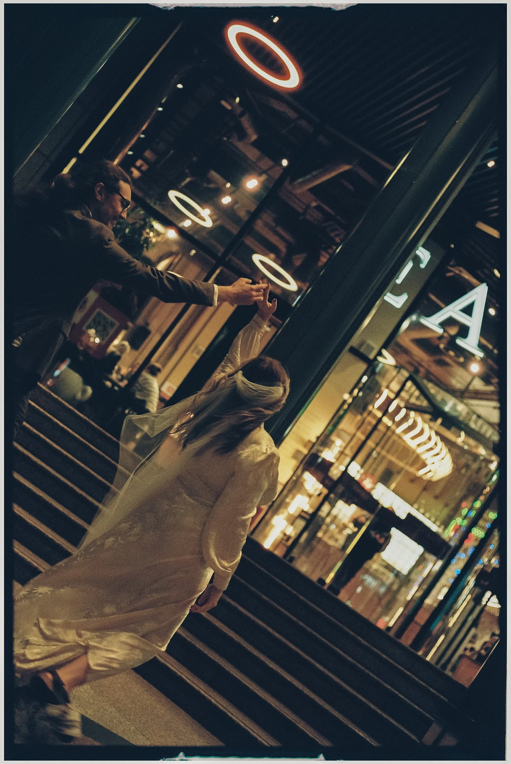 A man in a suit and a woman in a white dress holding hands and dancing outside a modern building with neon ring lights at night.