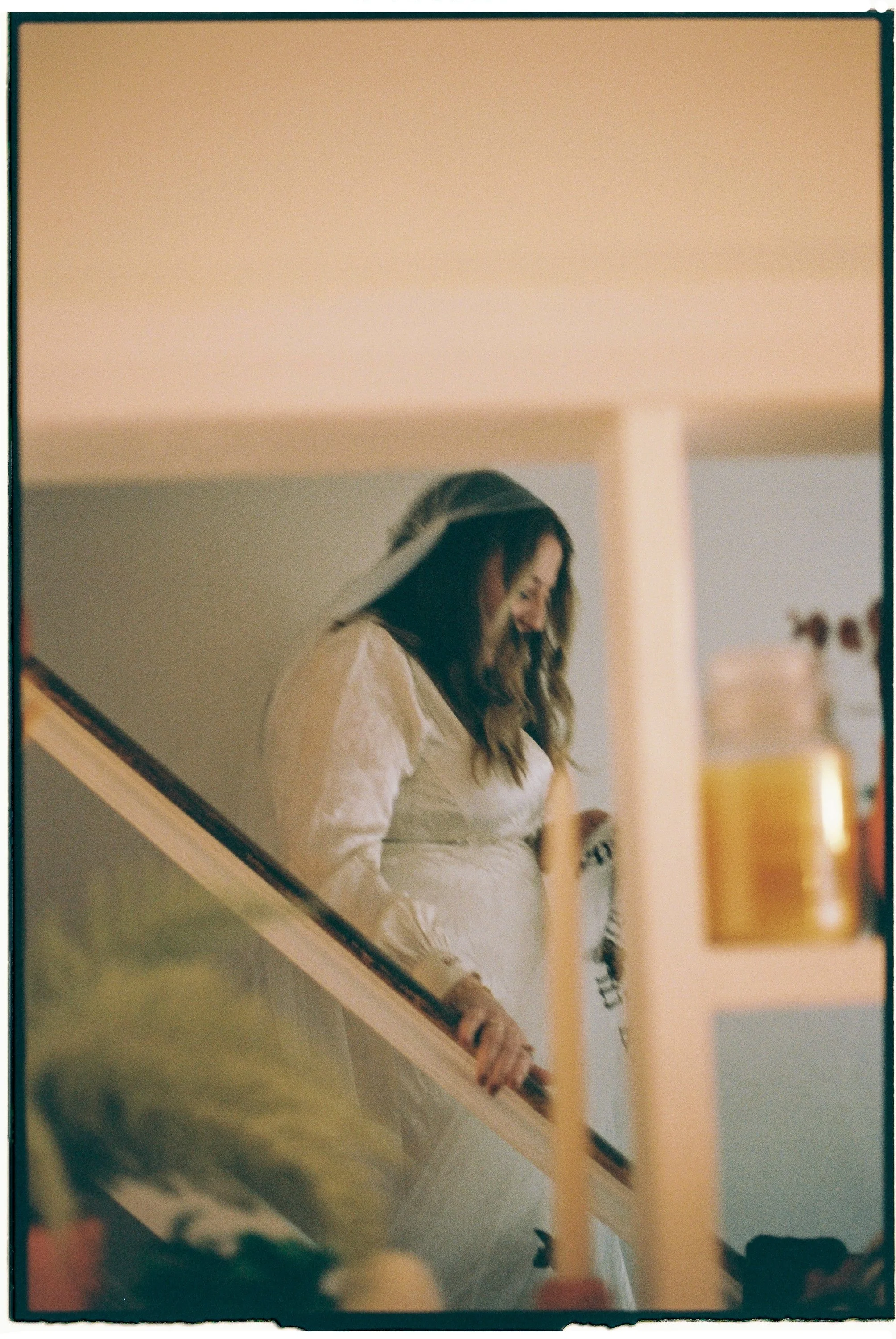 A woman in a white dress with lace details descending a staircase, smiling and looking down.