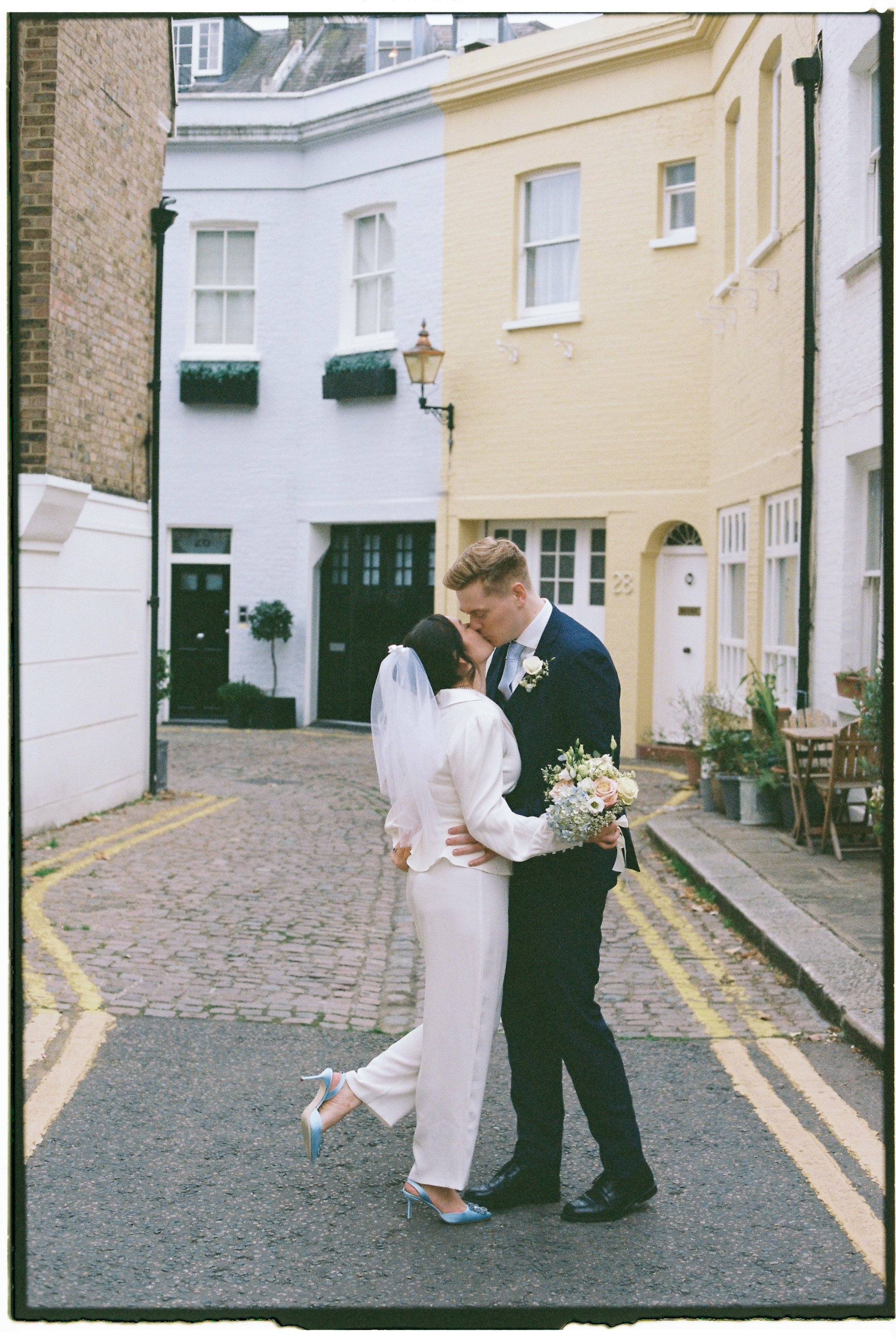 A newlywed couple kissing in a cobblestone alleyway, with the bride wearing a white pantsuit and veil, and the groom in a dark suit holding a bouquet of white and pink flowers.