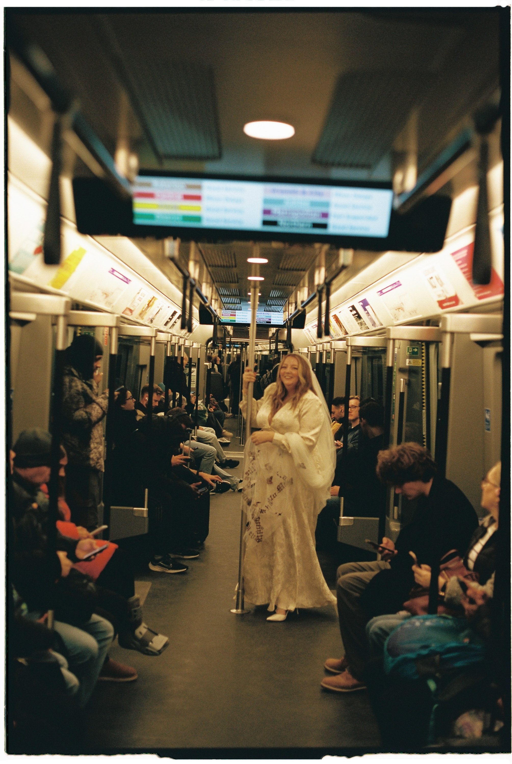 A woman dressed in a white, flowing gown standing inside a train, holding a pole, surrounded by seated passengers on both sides who are looking at her.