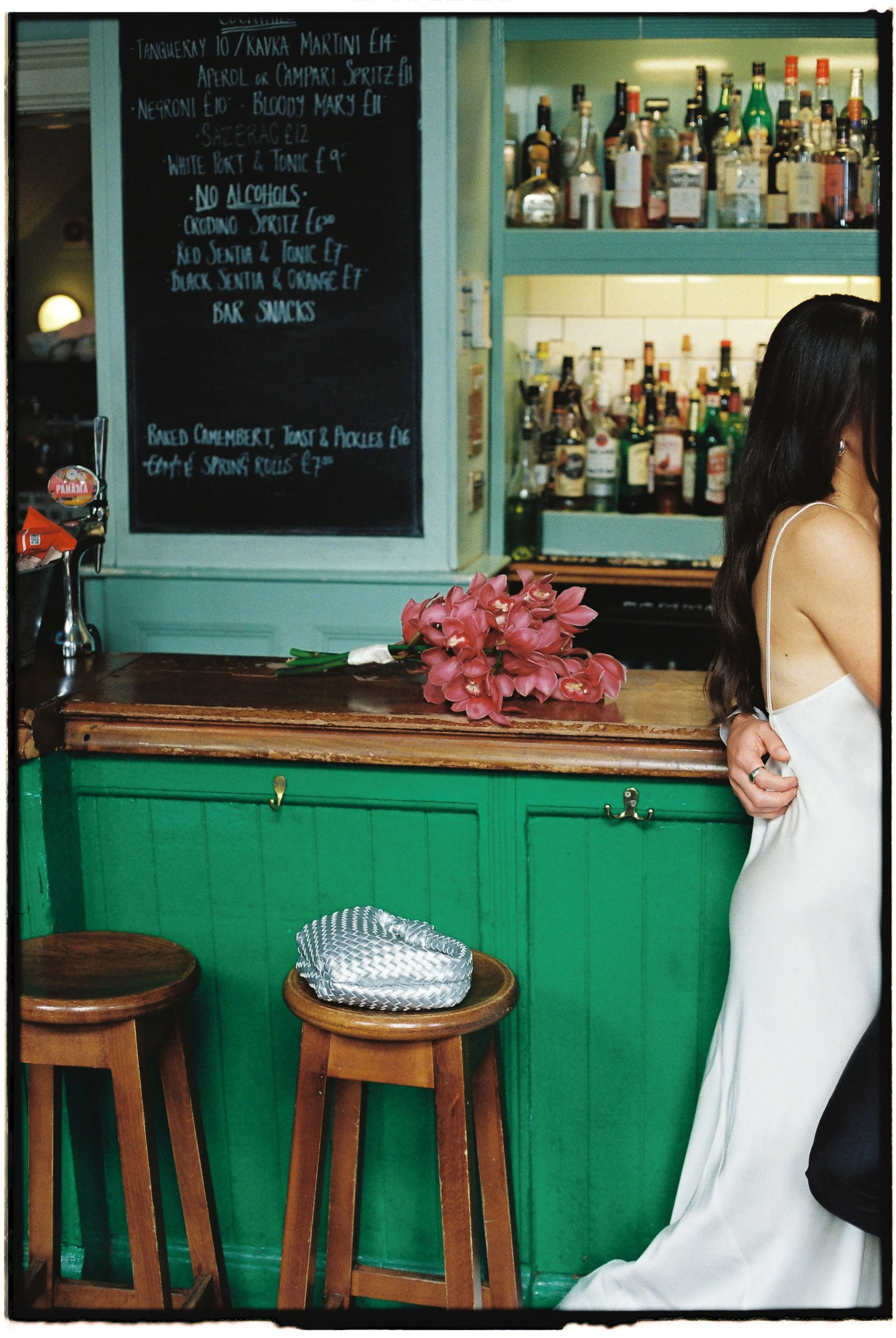 A bar with a green wooden counter, two wooden stools, and a bunch of pink flowers on the counter. Behind the bar, there are shelves stocked with various bottles of alcohol. A blackboard menu lists drinks, bar snacks, and a dessert item. Part of a wom