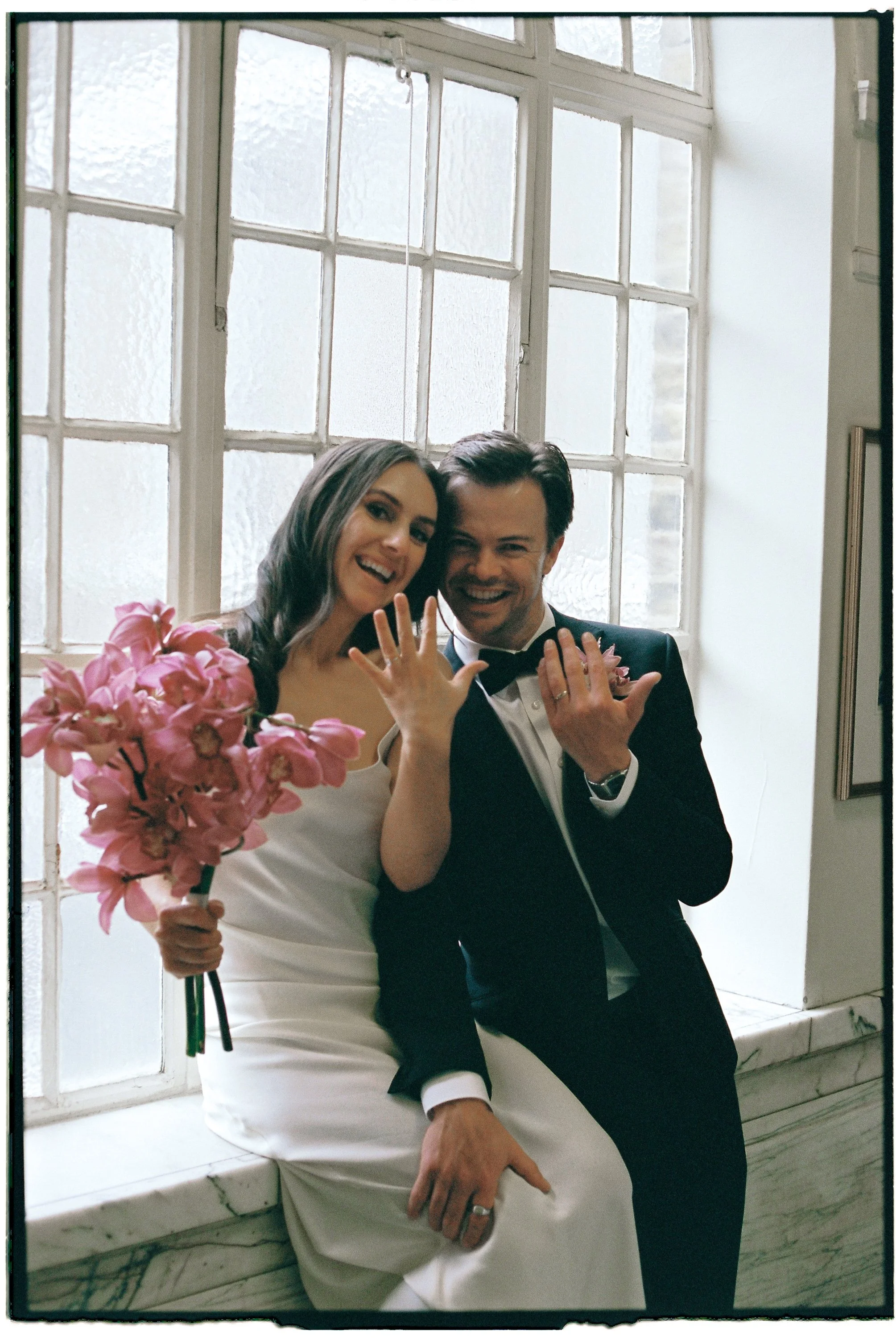 A newlywed couple posing near a large window, showing their wedding rings and the bride holding a bouquet of pink flowers.