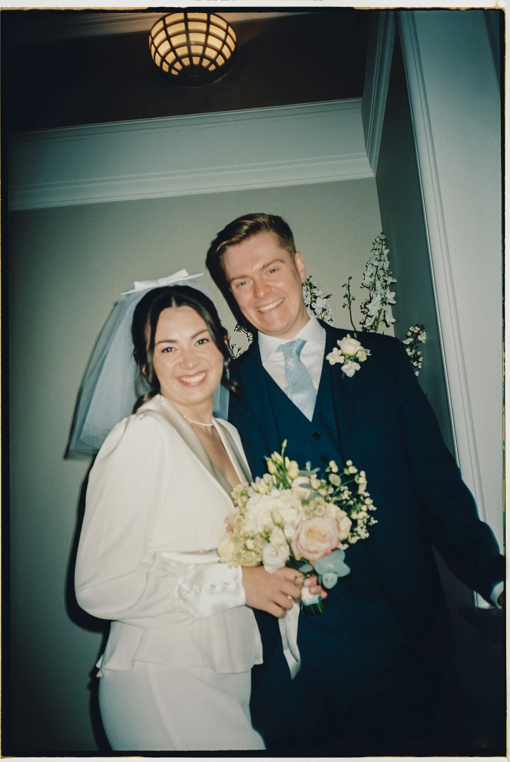 Young bride and groom smiling, standing indoors with flowers and greenery in the background, celebrating their wedding.