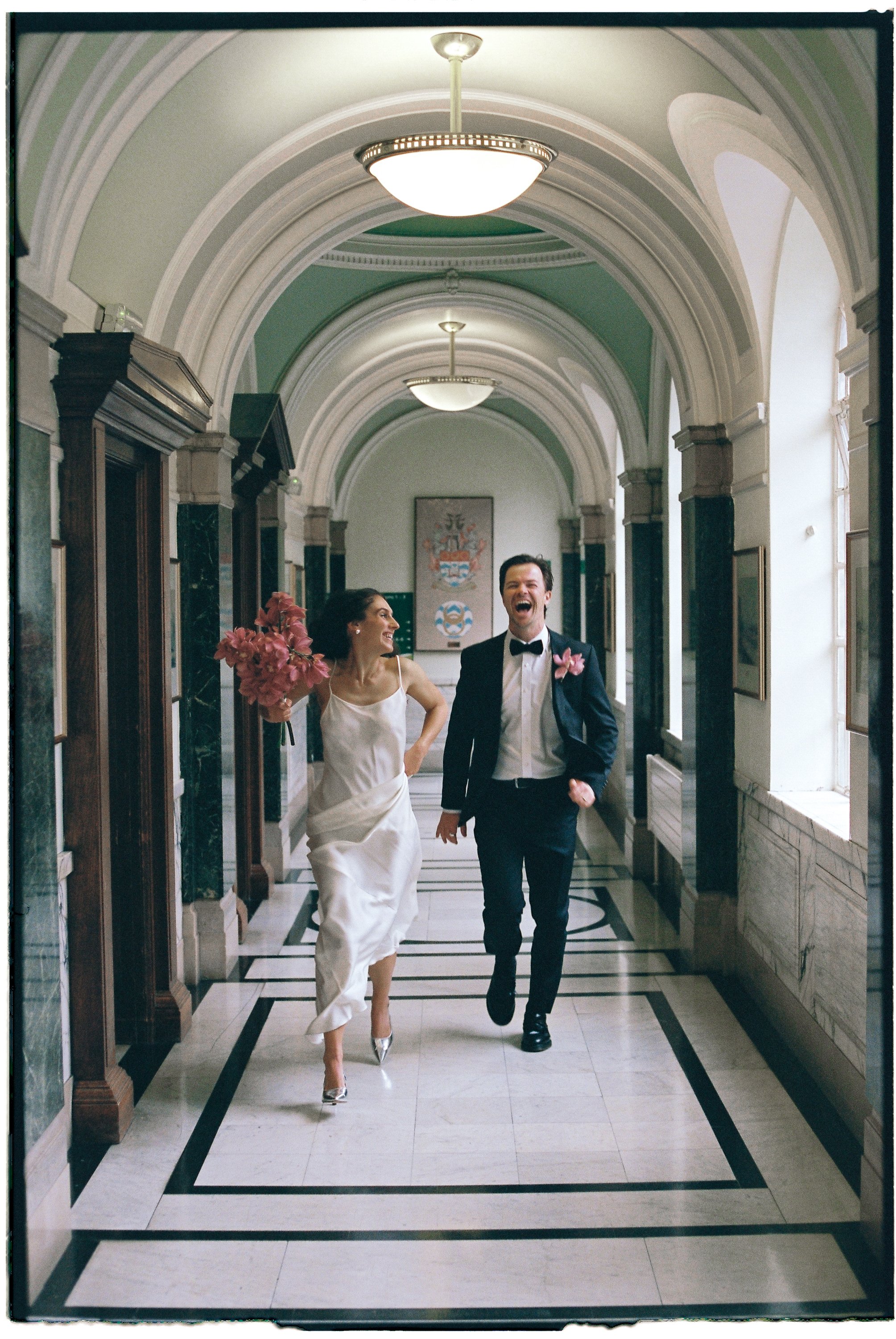 A bride in a white dress holding pink flowers and a groom in a tuxedo, both happy and laughing, walking through an elegant hallway with arched ceilings, chandeliers, and artwork on the walls.