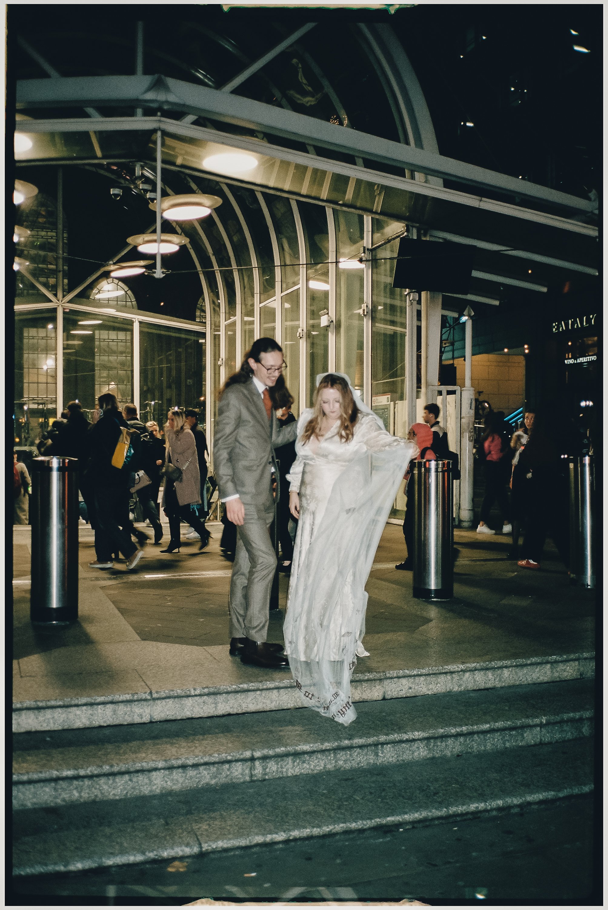 A couple dressed in wedding attire walking down the steps outside a modern glass building at night, with several people in the background.