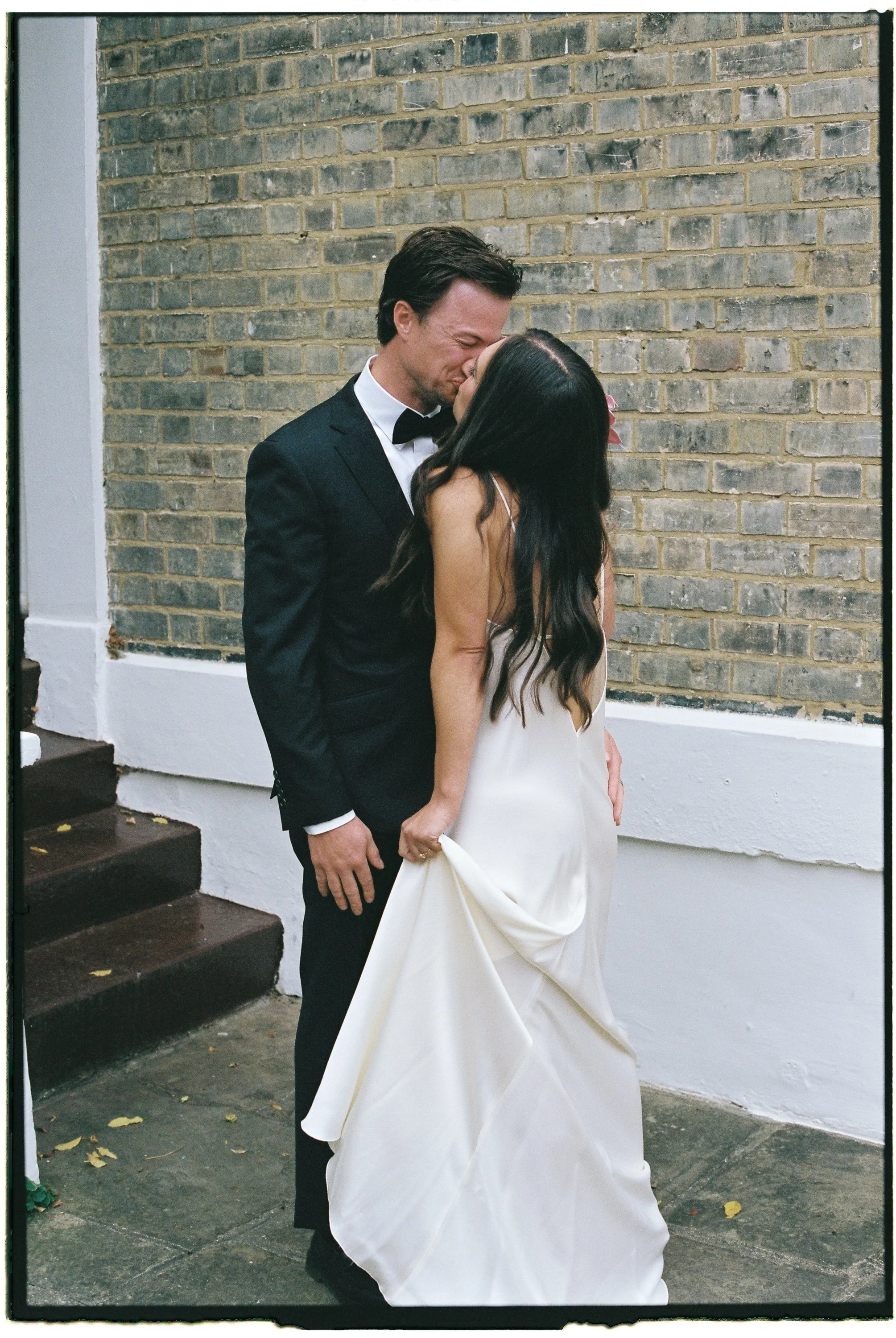 A couple in wedding attire sharing a kiss. The groom is wearing a black tuxedo with a bow tie, and the bride is in a white wedding gown with long dark hair. They are standing against a brick wall near some steps.