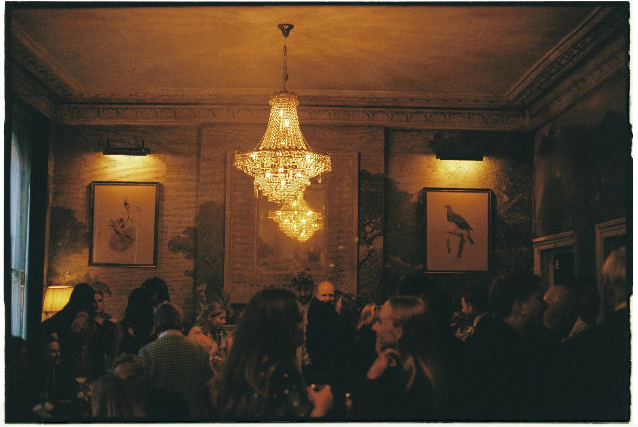 An elegant dining room filled with people, illuminated by a large crystal chandelier hanging from a decorated ceiling, with framed artwork and wall lamps.