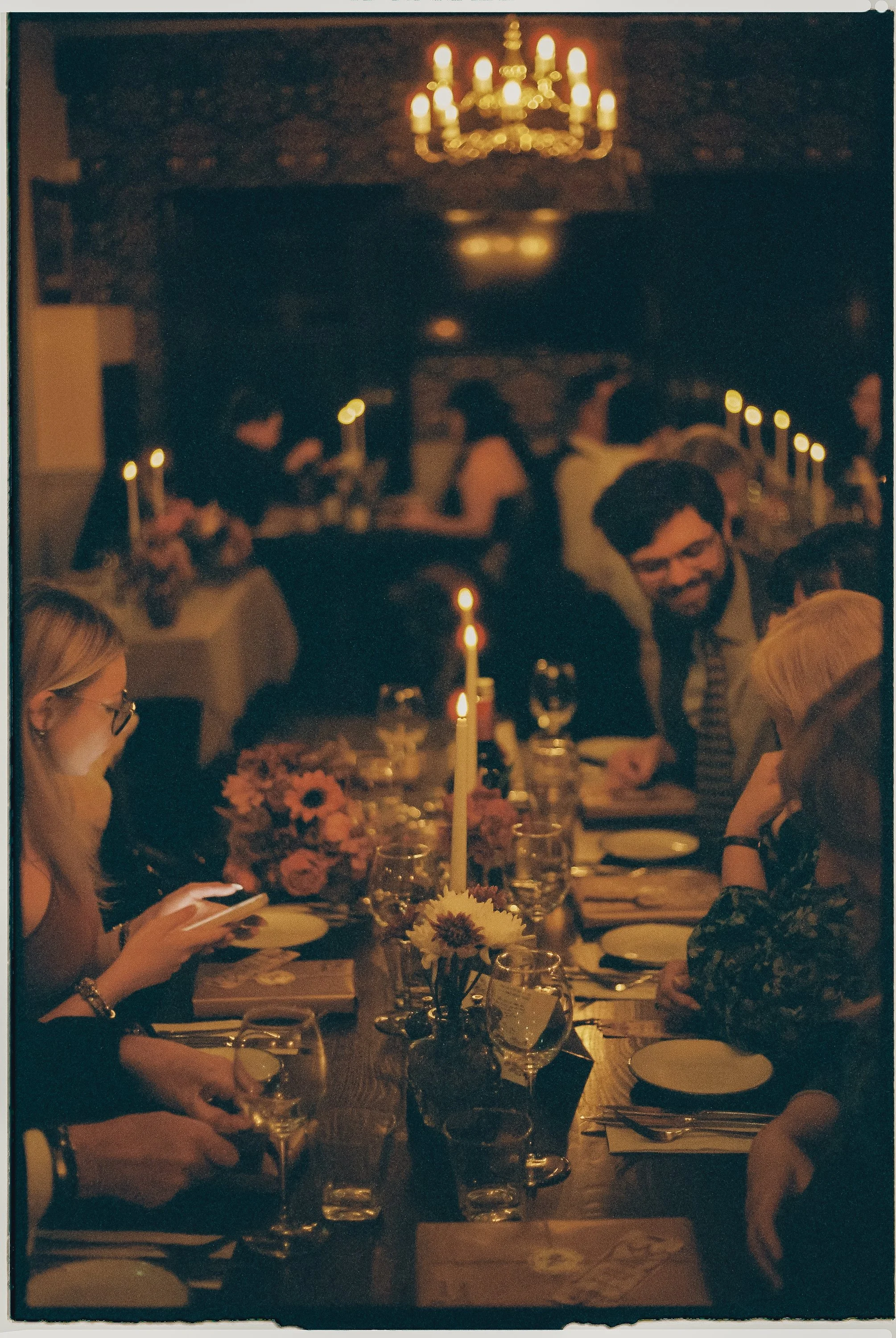 People dining at a candlelit dinner party in a dimly lit room with floral centerpieces and a chandelier above.