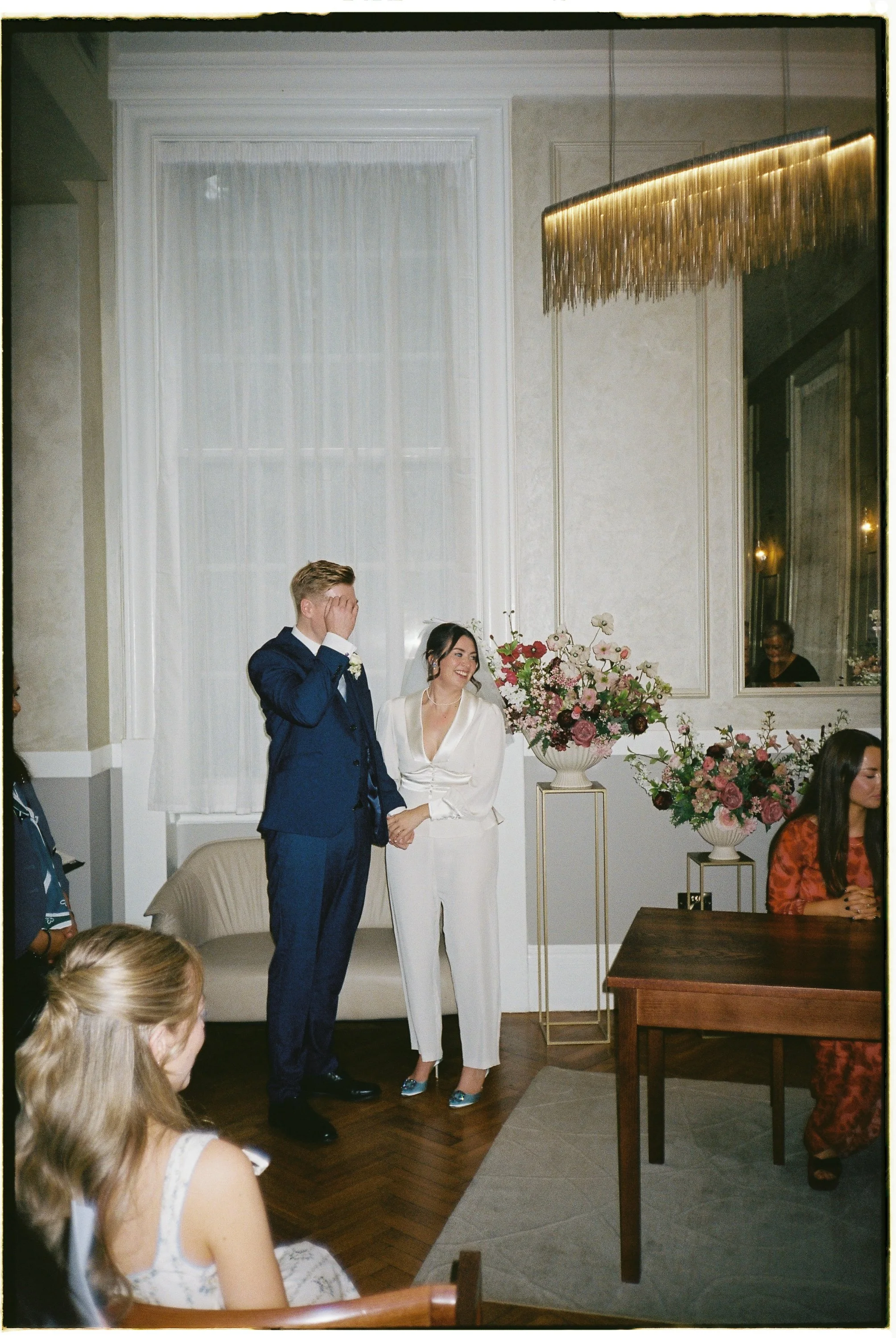 A wedding ceremony with a bride and groom standing hand in hand; the groom covers his face with his hand, and the bride is smiling. There are floral arrangements next to them, and guests seated around the room.
