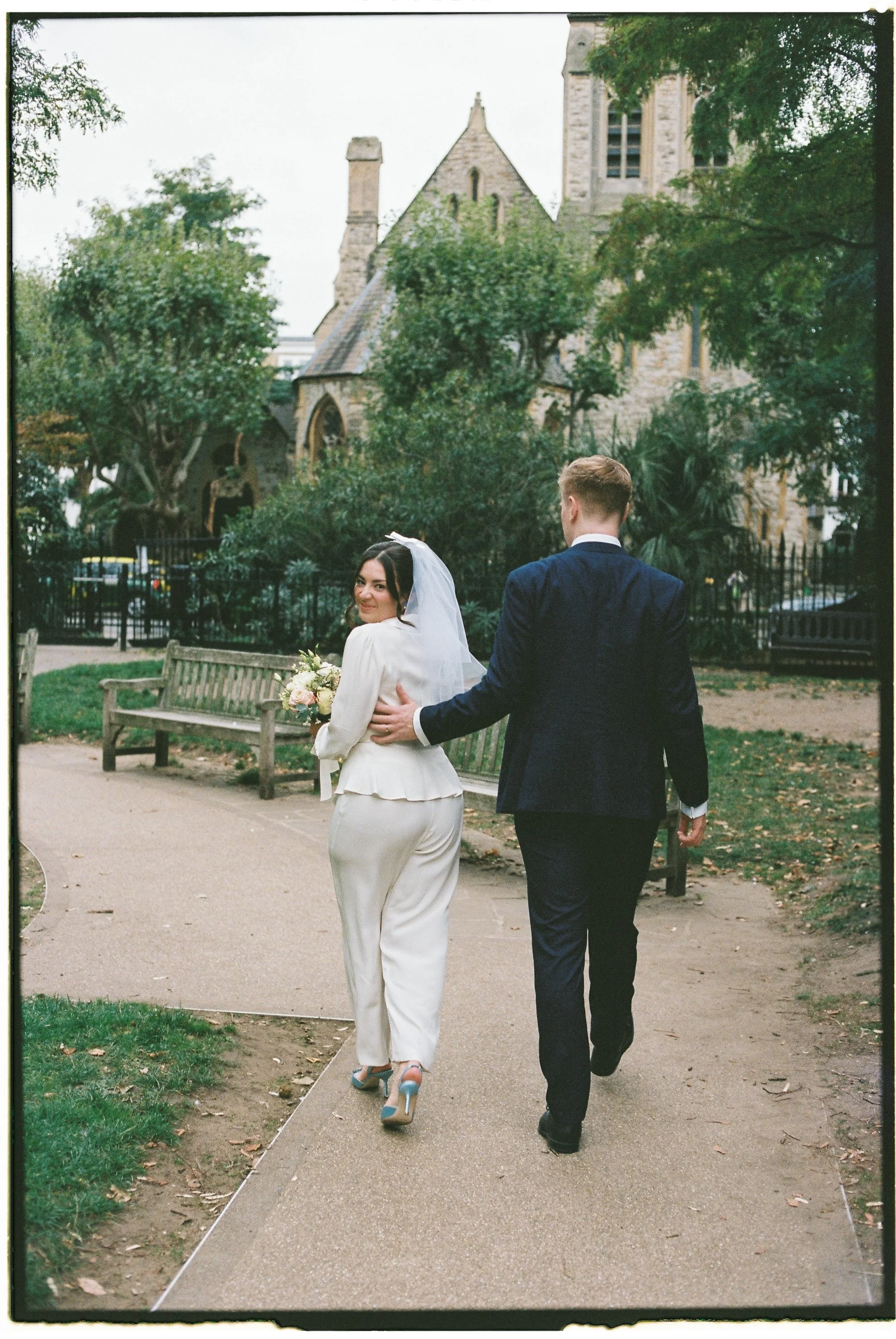 A bride and groom walking in a park, with the bride smiling at the camera and holding a bouquet, and the groom behind her gently touching her back, with a historic stone church and greenery in the background.