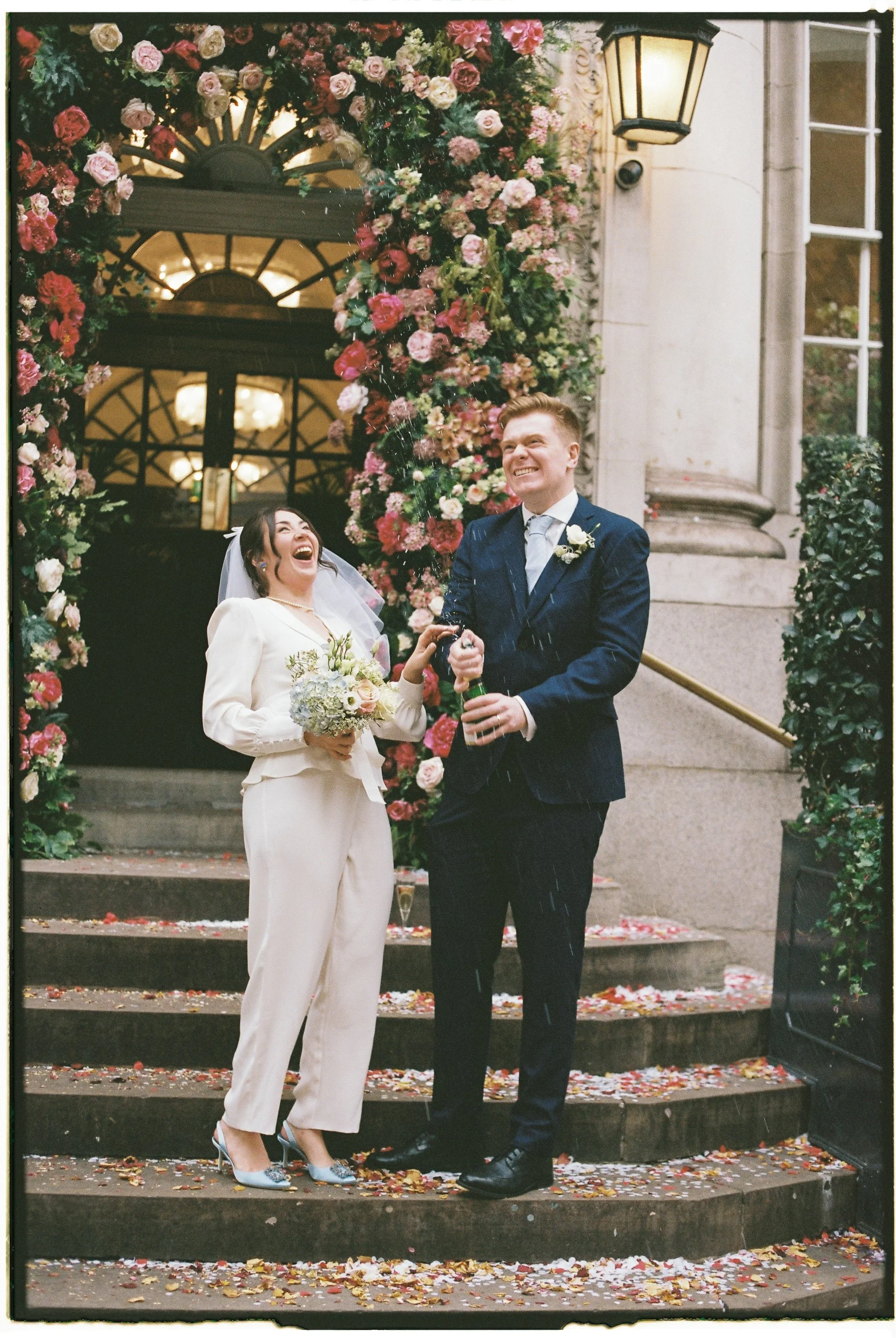 A bride and groom celebrating their wedding on the steps outside a building decorated with a large arch of pink and white flowers, with confetti and flower petals on the ground, and a lantern hanging on the wall.