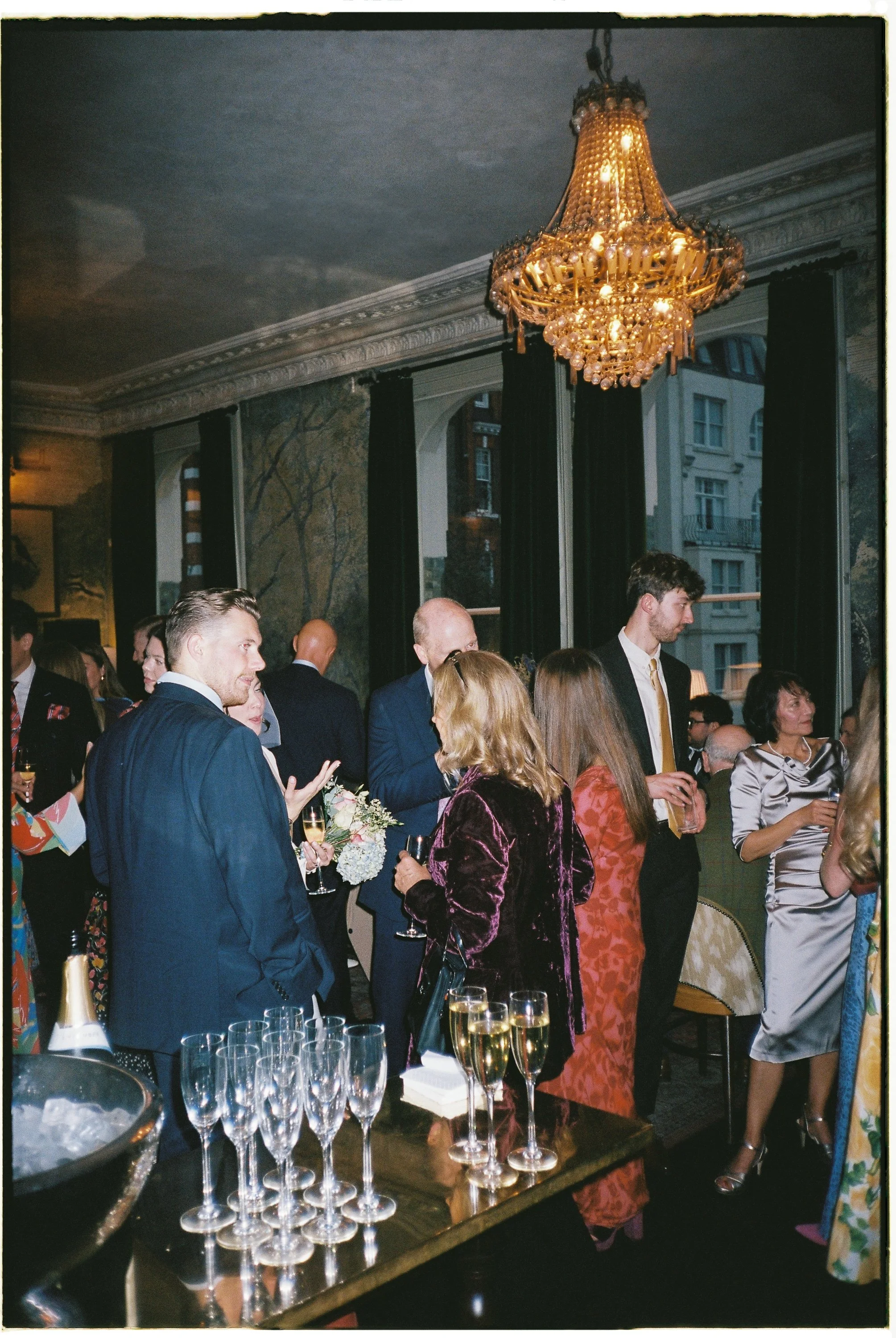 People socializing at an upscale indoor event with a chandelier hanging from the ceiling, dark curtains, and large windows showing buildings outside.