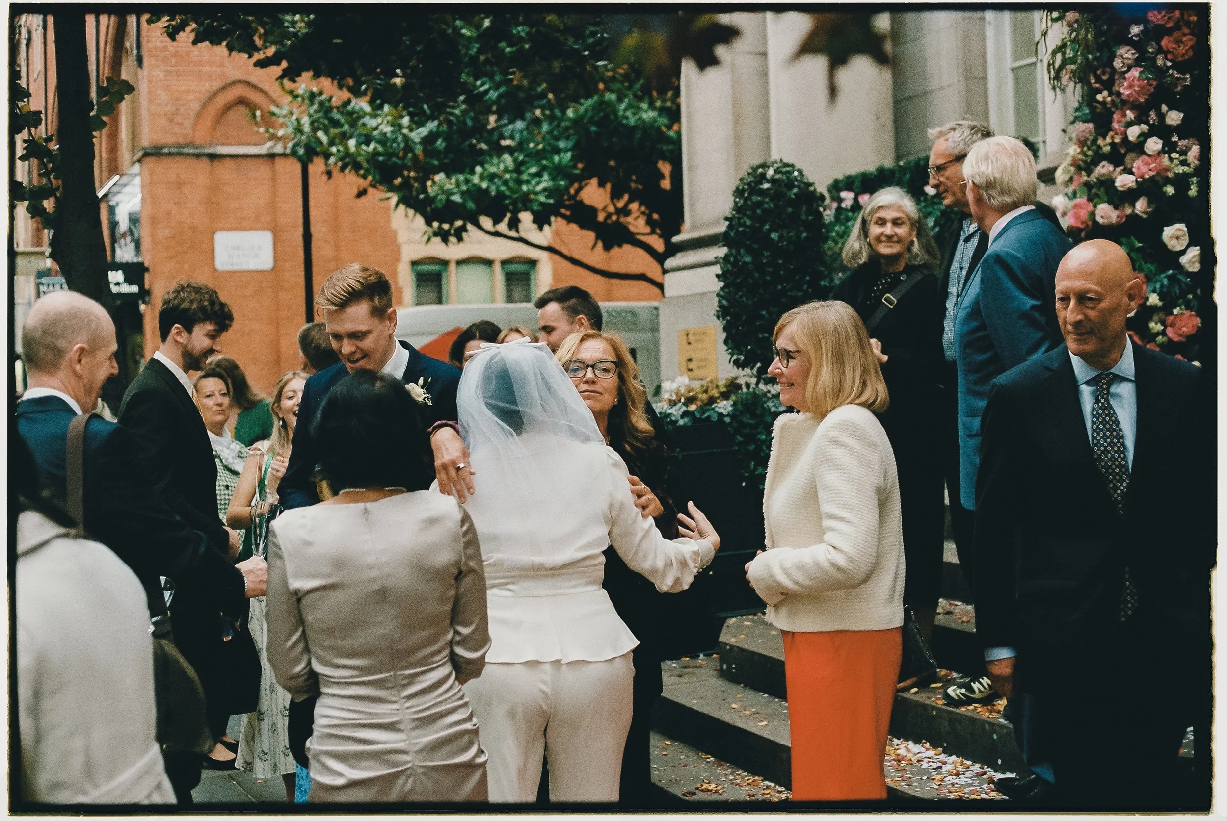 People gathered outside a building at a wedding celebration, with some embracing and smiling, others chatting and standing around, and flowers decorating the area.