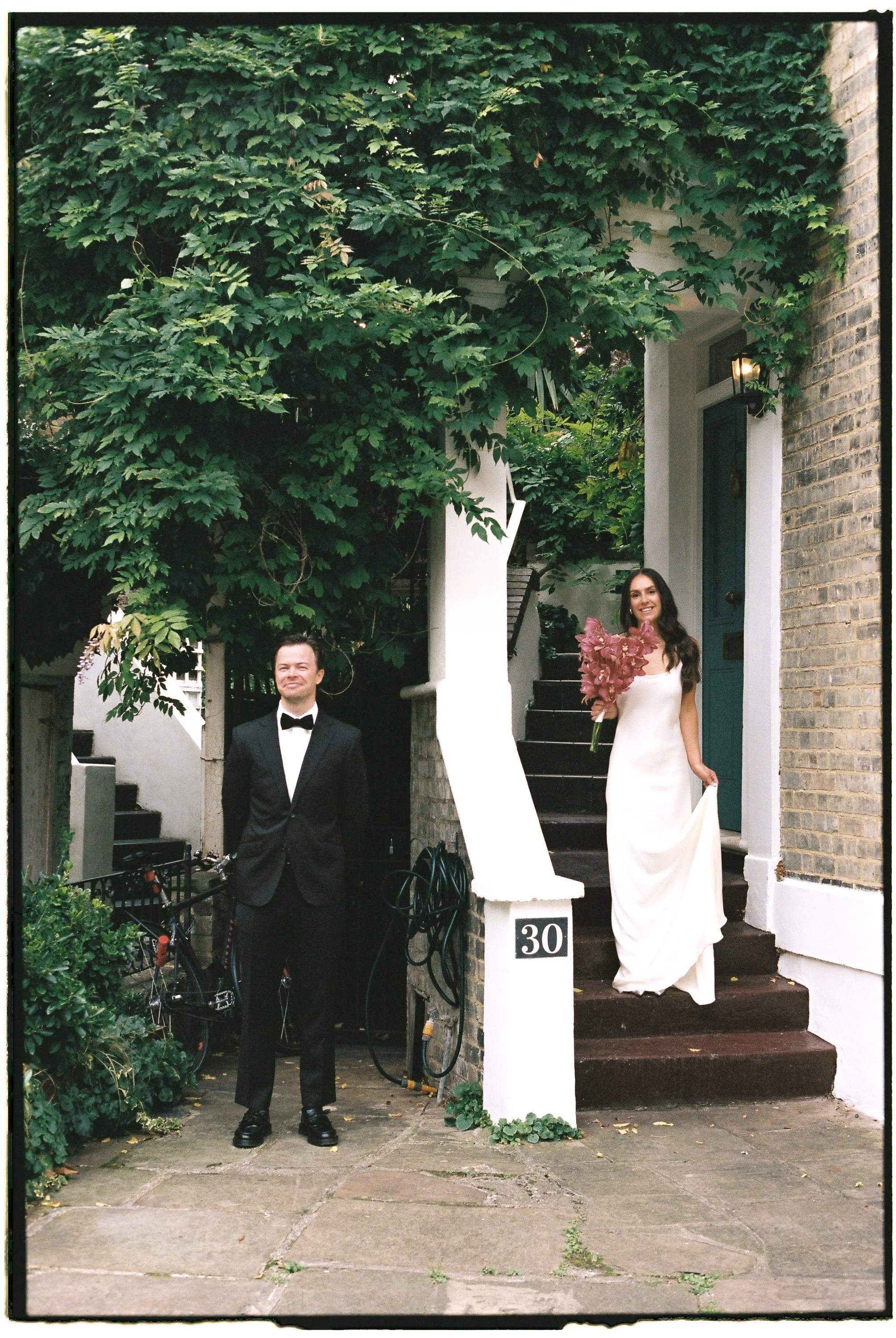 A man and a woman dressed in formal attire are standing outside a house. The man is in a tuxedo with a bow tie, and the woman is in a white dress holding a bouquet of pink flowers. The woman stands on the steps of the house by the door, while the man