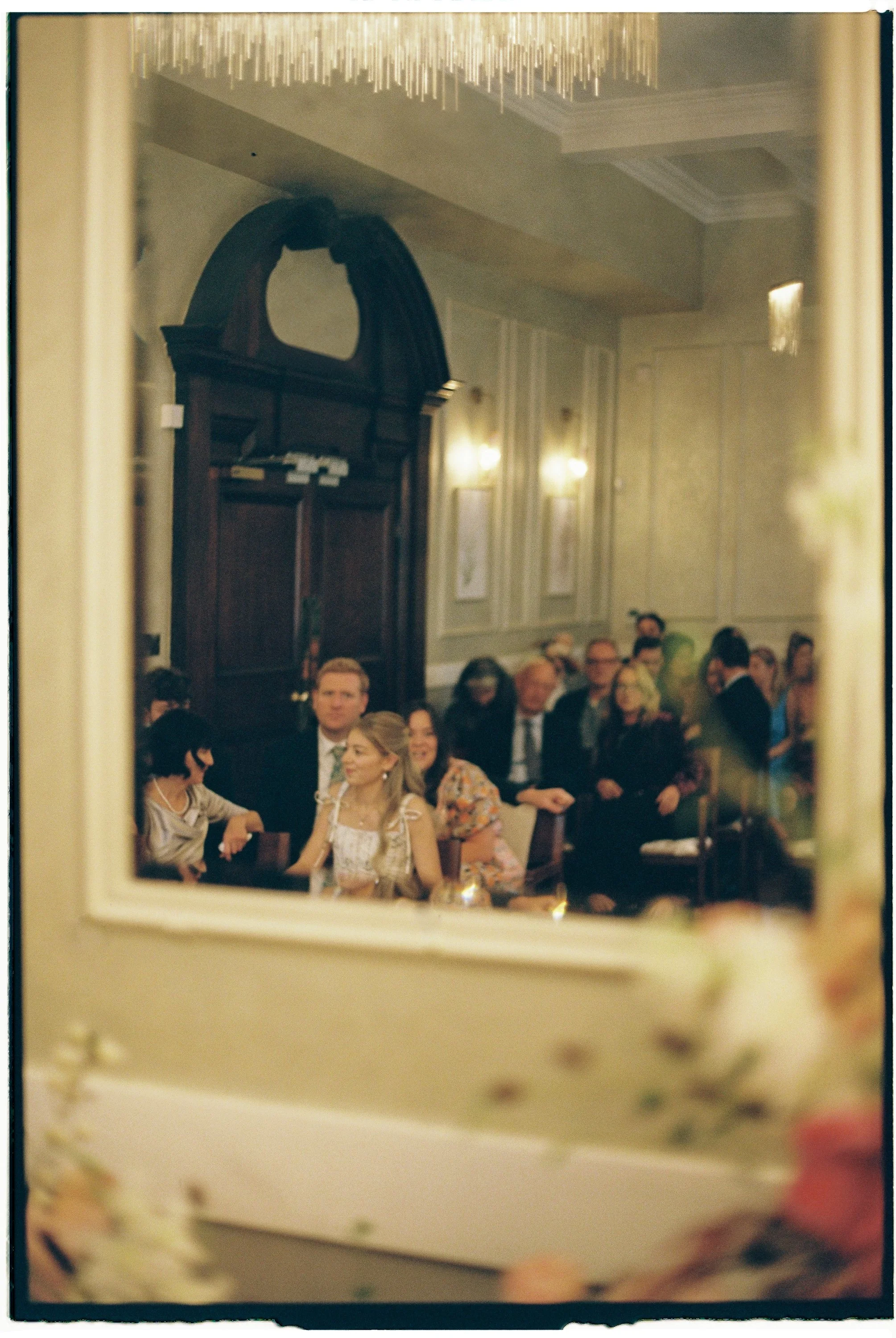 People attending a formal event or celebration in a banquet hall, seen through a mirror with floral decorations at the bottom of the frame.