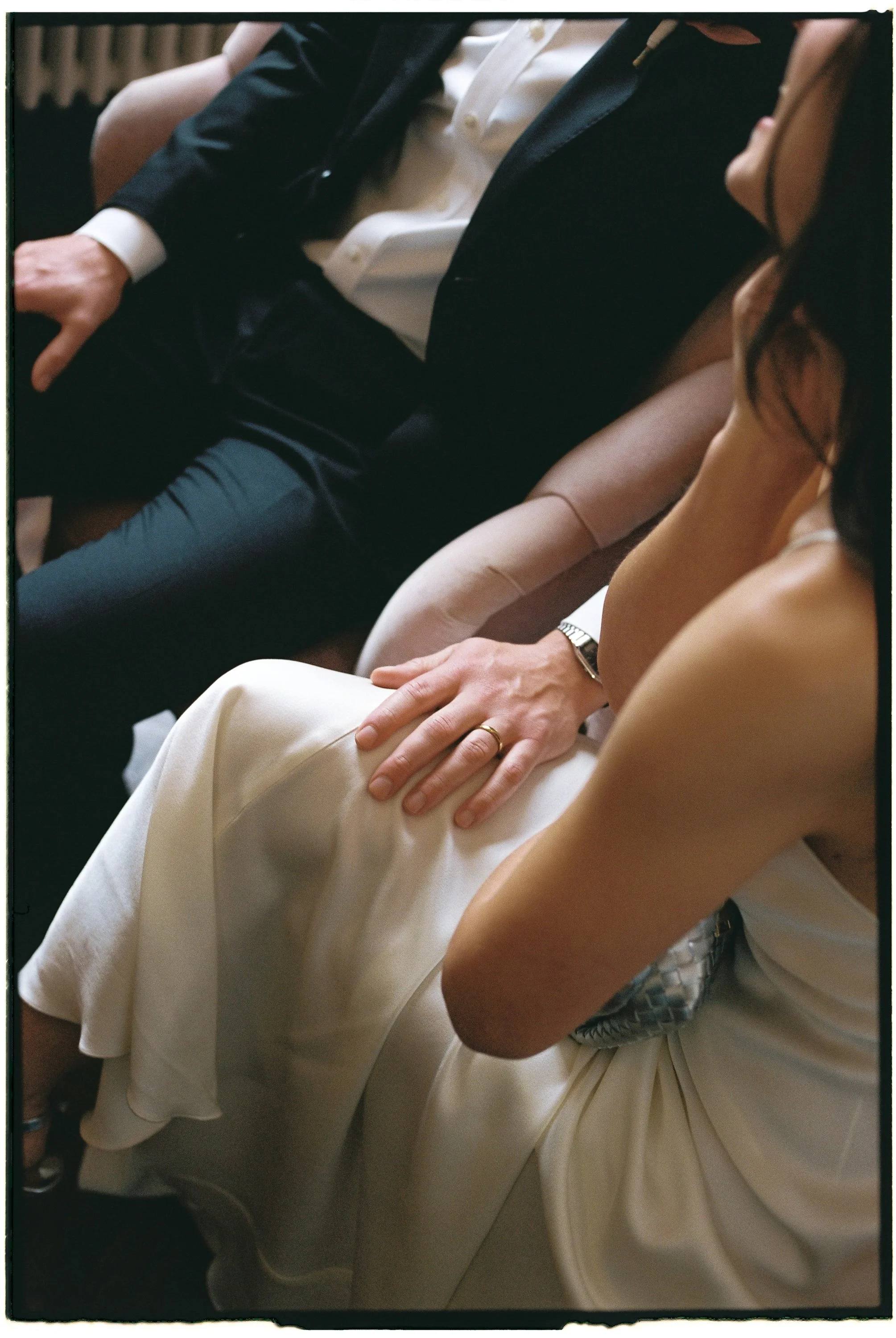 Close-up of a wedding ceremony scene with a bride and groom seated, the bride's hand resting on her lap, showing a wedding ring, and the groom's hand on his knee, dressed in formal attire.
