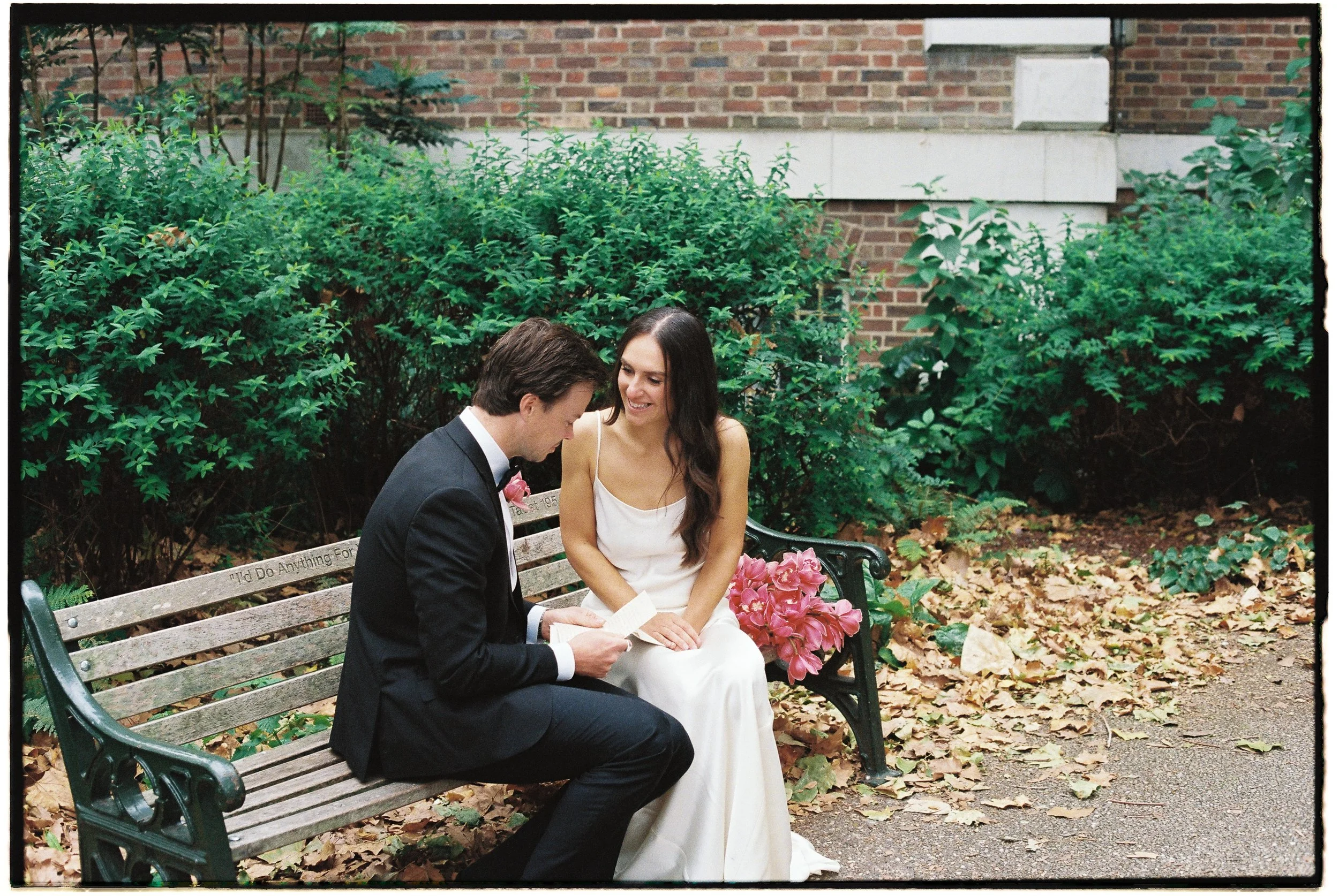 A man in a black suit and a woman in a white dress sit on a park bench with pink flowers, surrounded by bushes and fallen leaves, in front of a brick building.