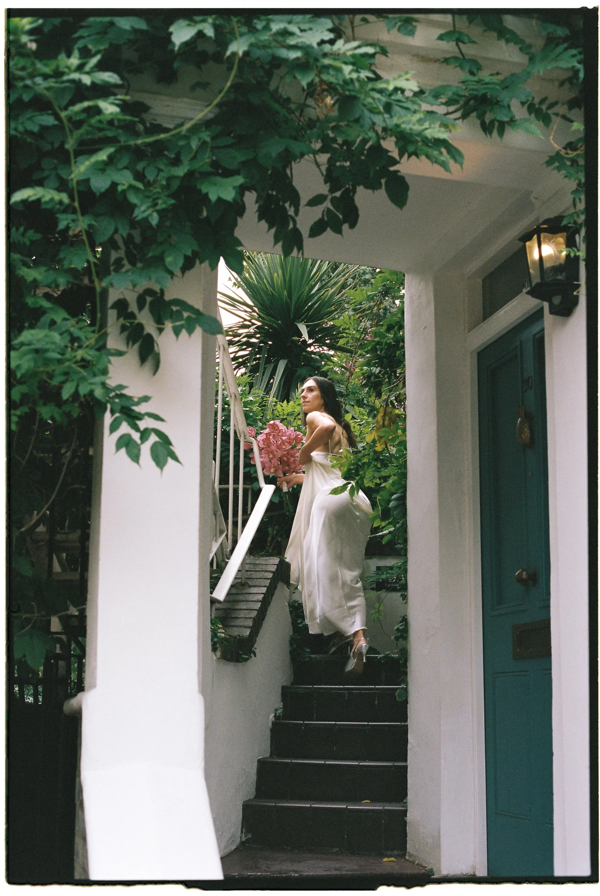 A woman in a white satin gown standing on stairs surrounded by green plants and trees, holding a bouquet of pink flowers, near a teal door with a black lantern.