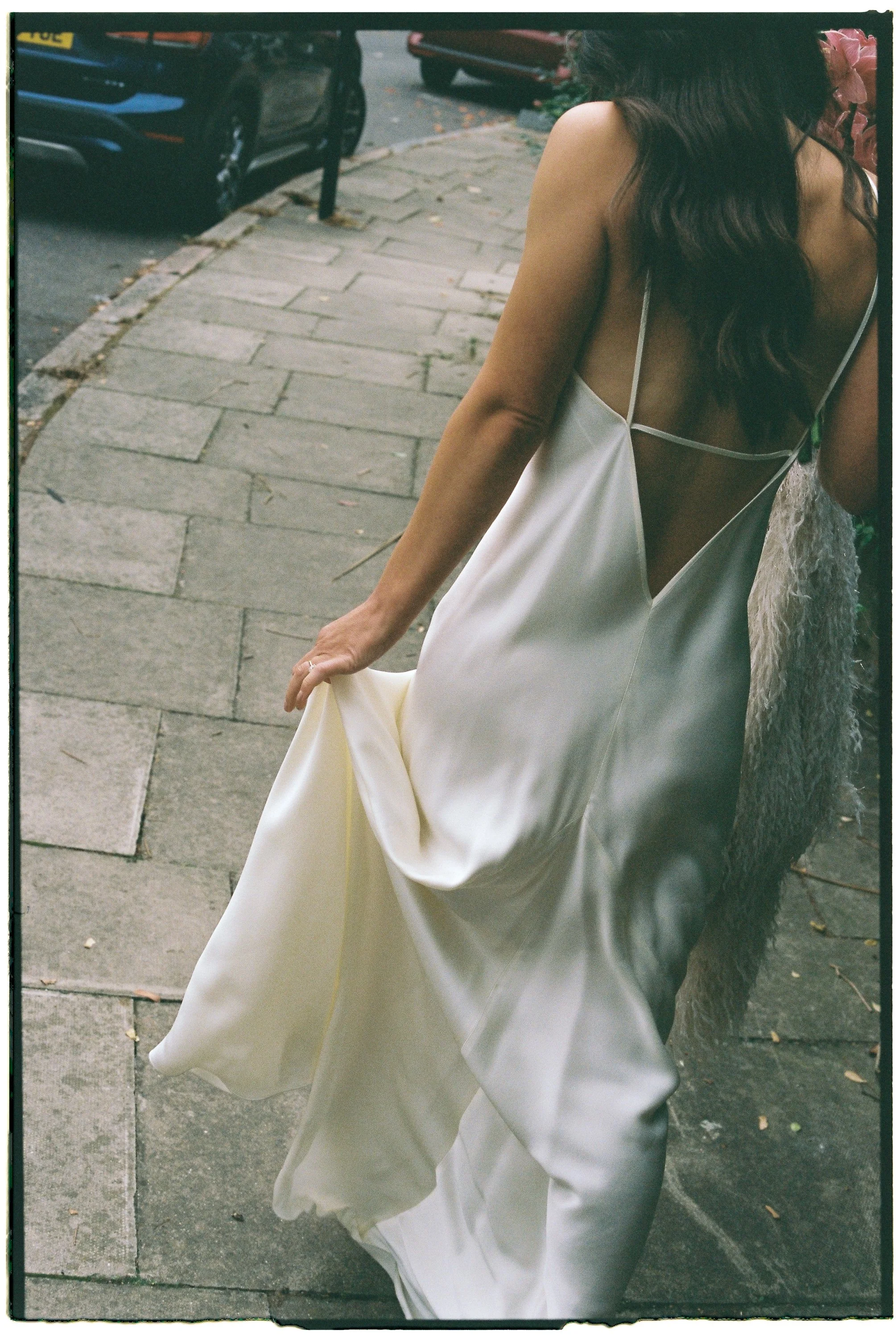 A woman in an elegant white satin gown with thin straps and an open back is standing on a sidewalk, lifting the hem of her dress slightly.
