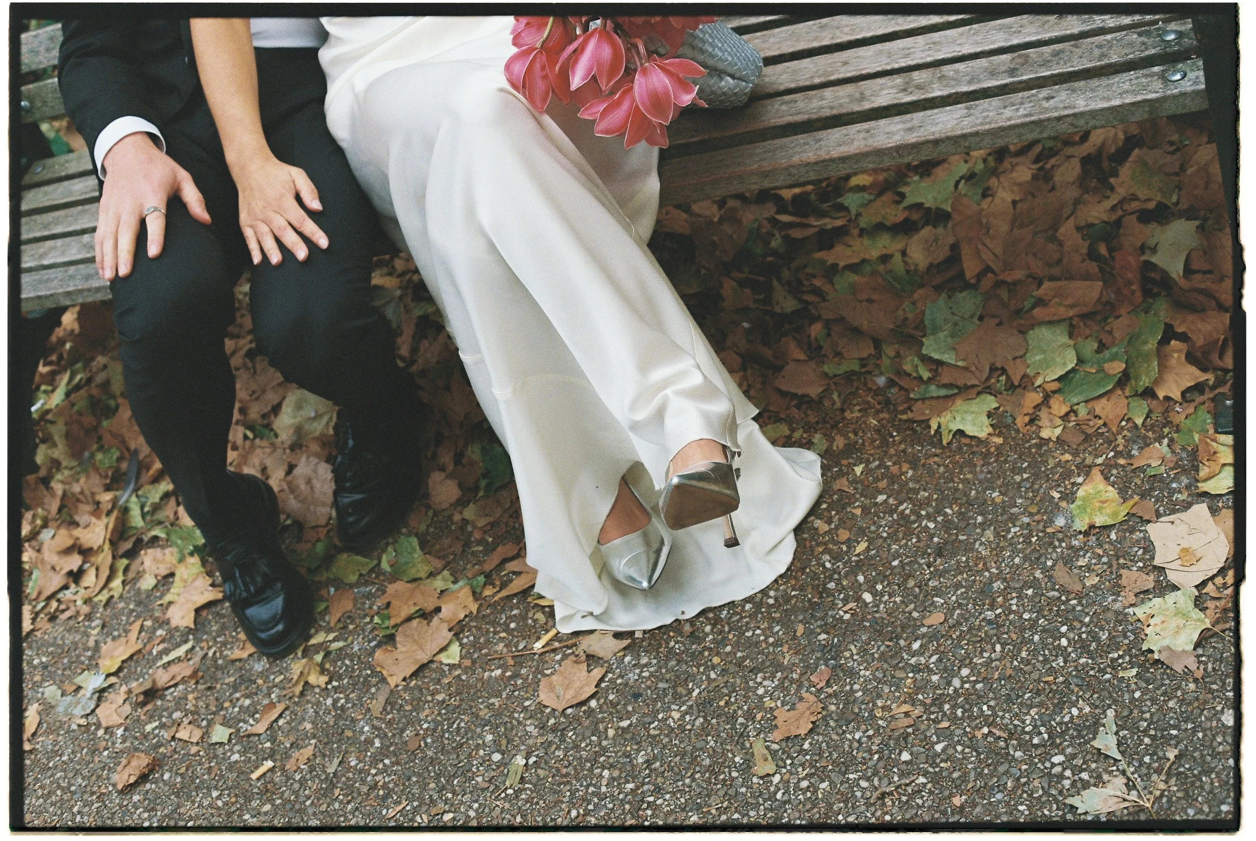 Couple sitting on a park bench with fallen autumn leaves on the ground, the woman wearing a white dress and silver high heels, holding a bouquet of pink flowers, and the man in black shoes and pants.