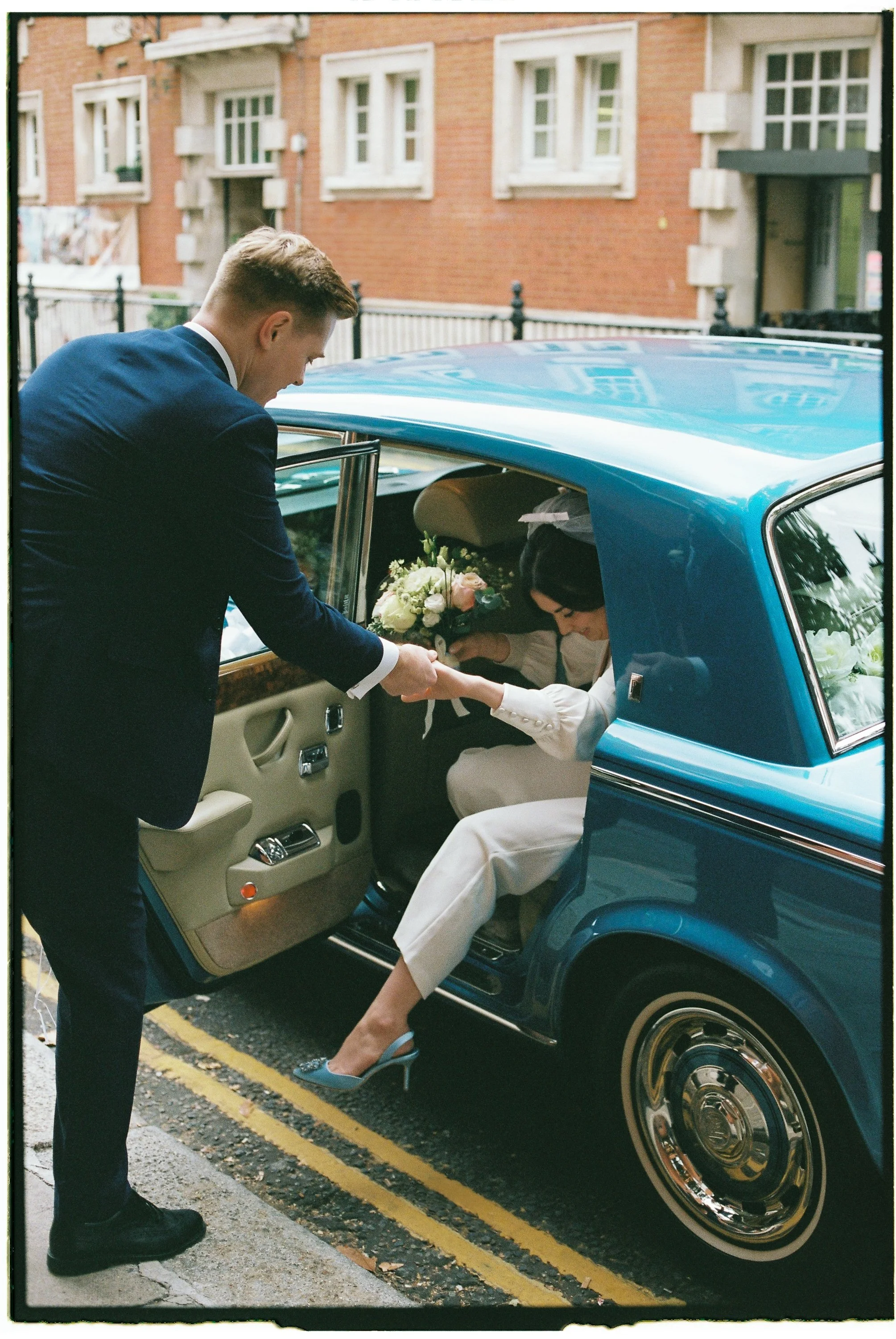 A man helping a woman into a vintage blue car on a city street, woman holding a bouquet of flowers, dressed in white, man in a dark suit.
