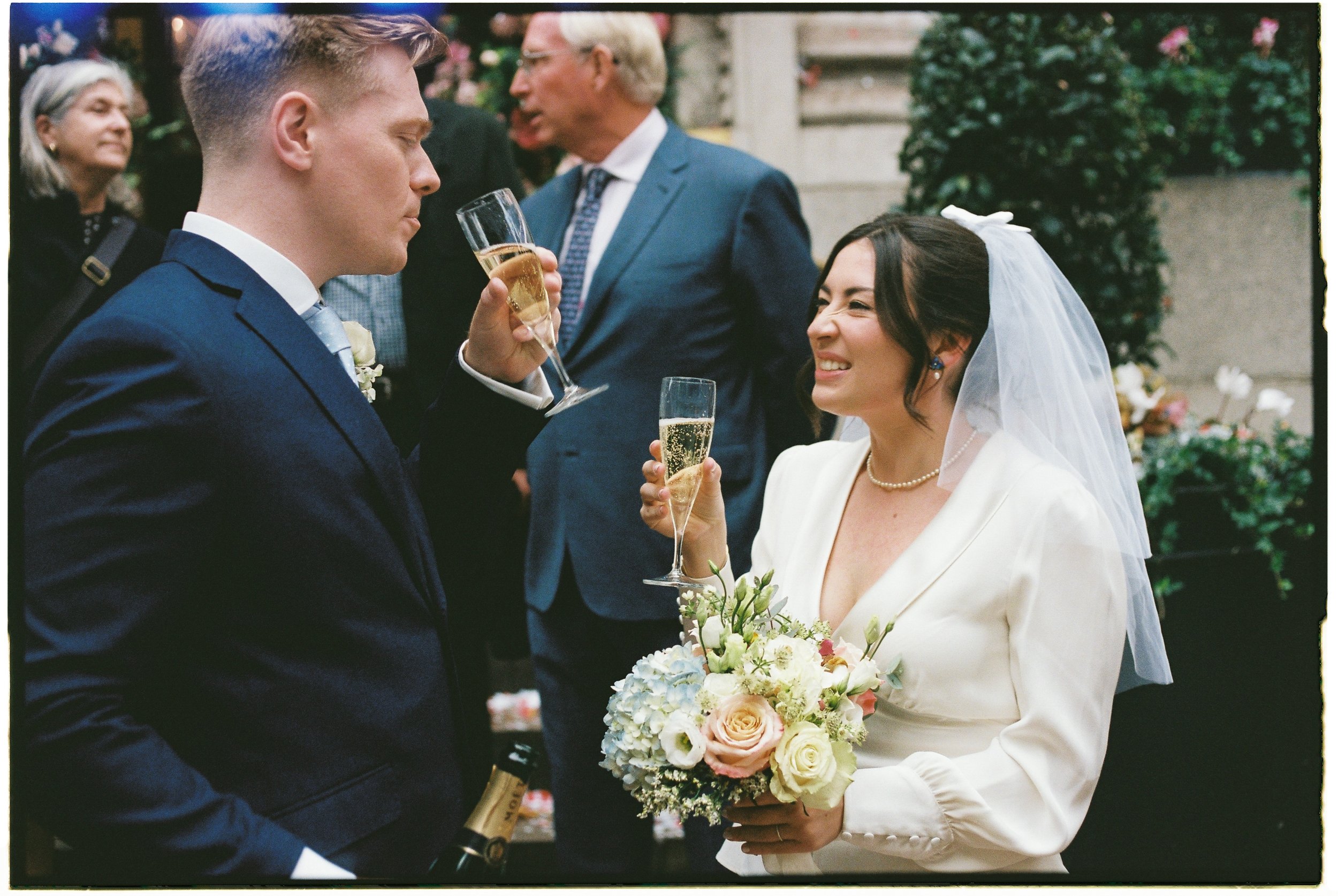 Bride and groom at wedding reception holding champagne glasses, with bride holding a bouquet of flowers, smiling and enjoying the celebration.