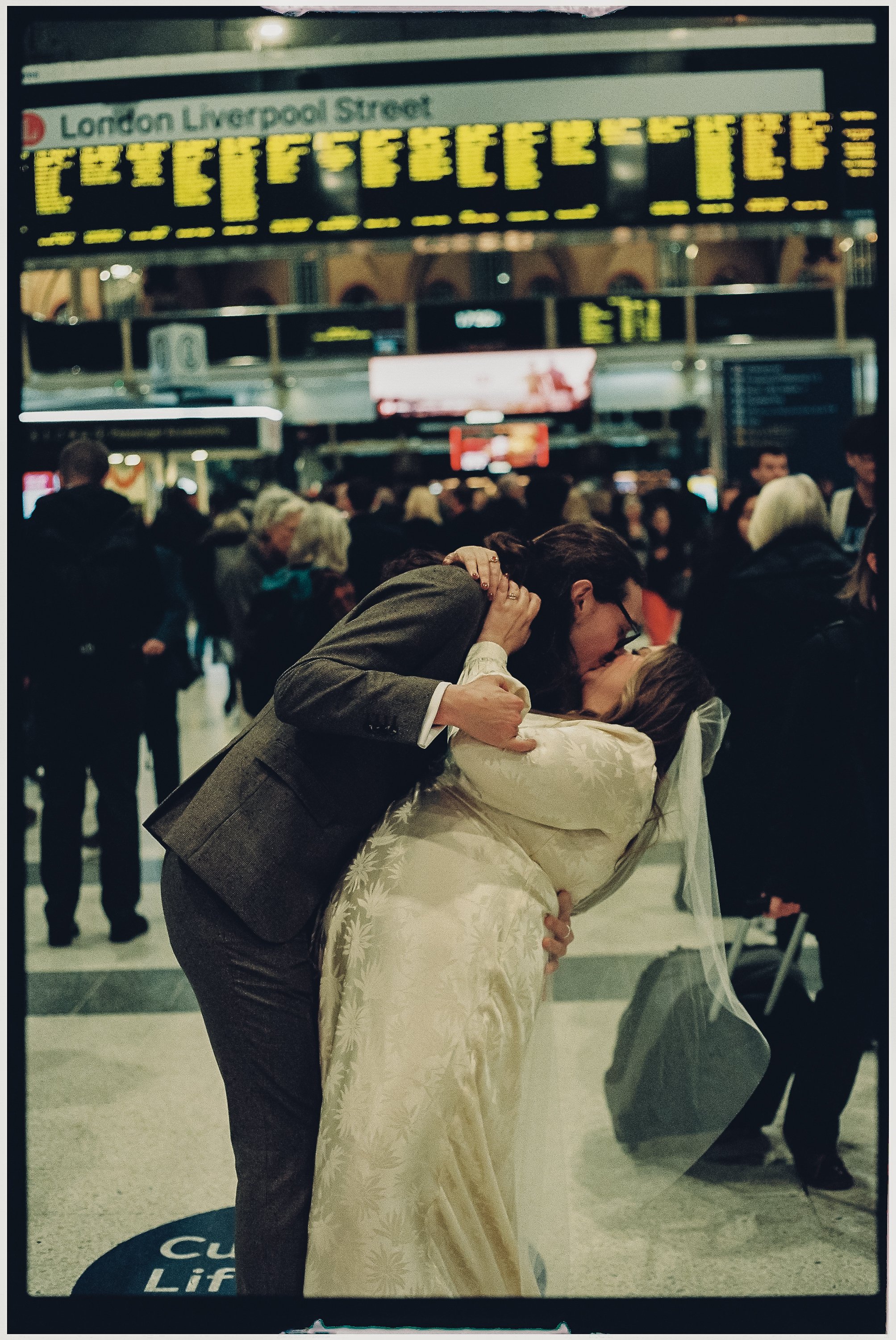 A couple kissing at an airport, with the woman in a wedding dress and the man in a suit, surrounded by busy travelers.