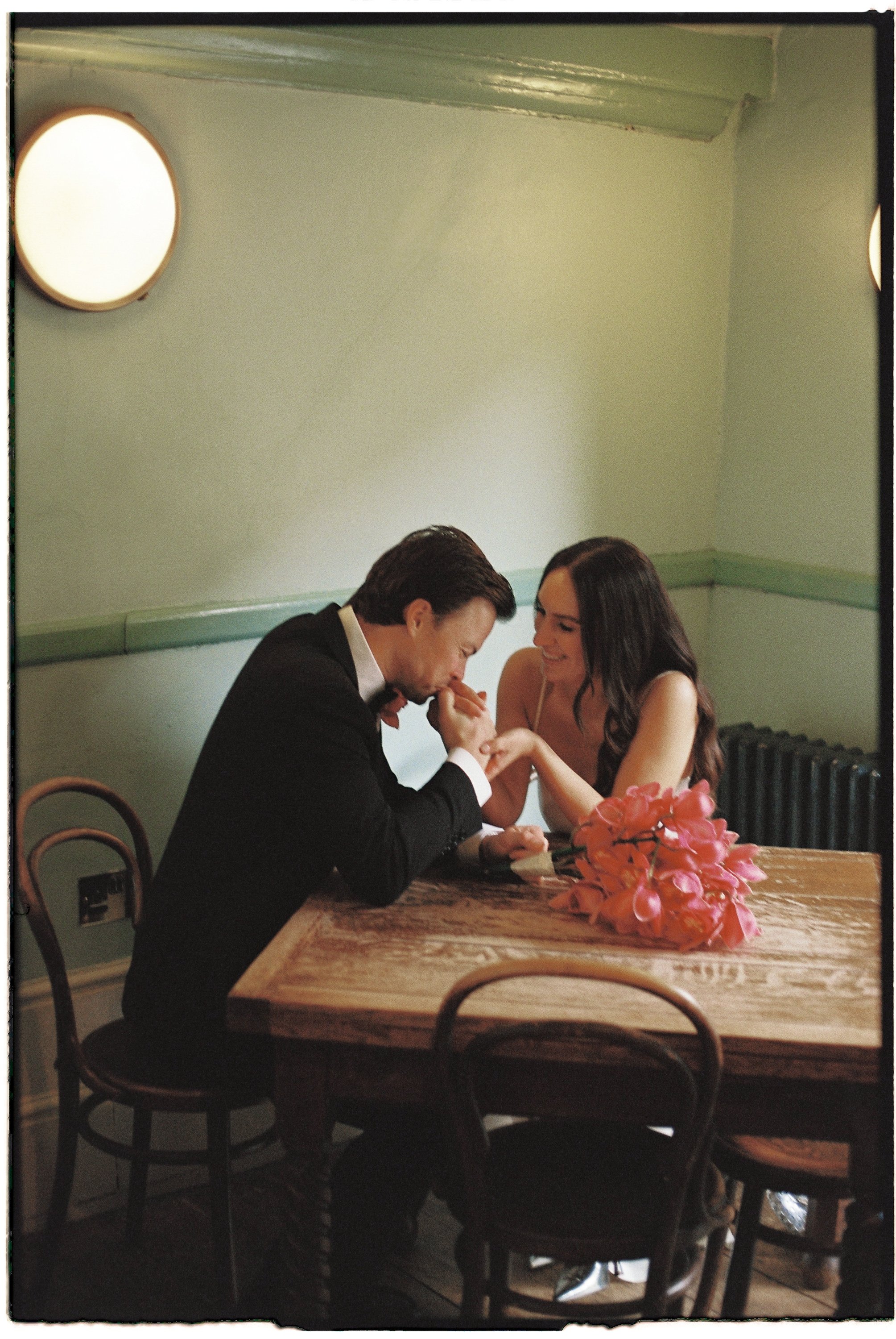 A man in a tuxedo and a woman in a white dress are sitting at a wooden table, engaging in a romantic moment, with a bouquet of pink flowers on the table.