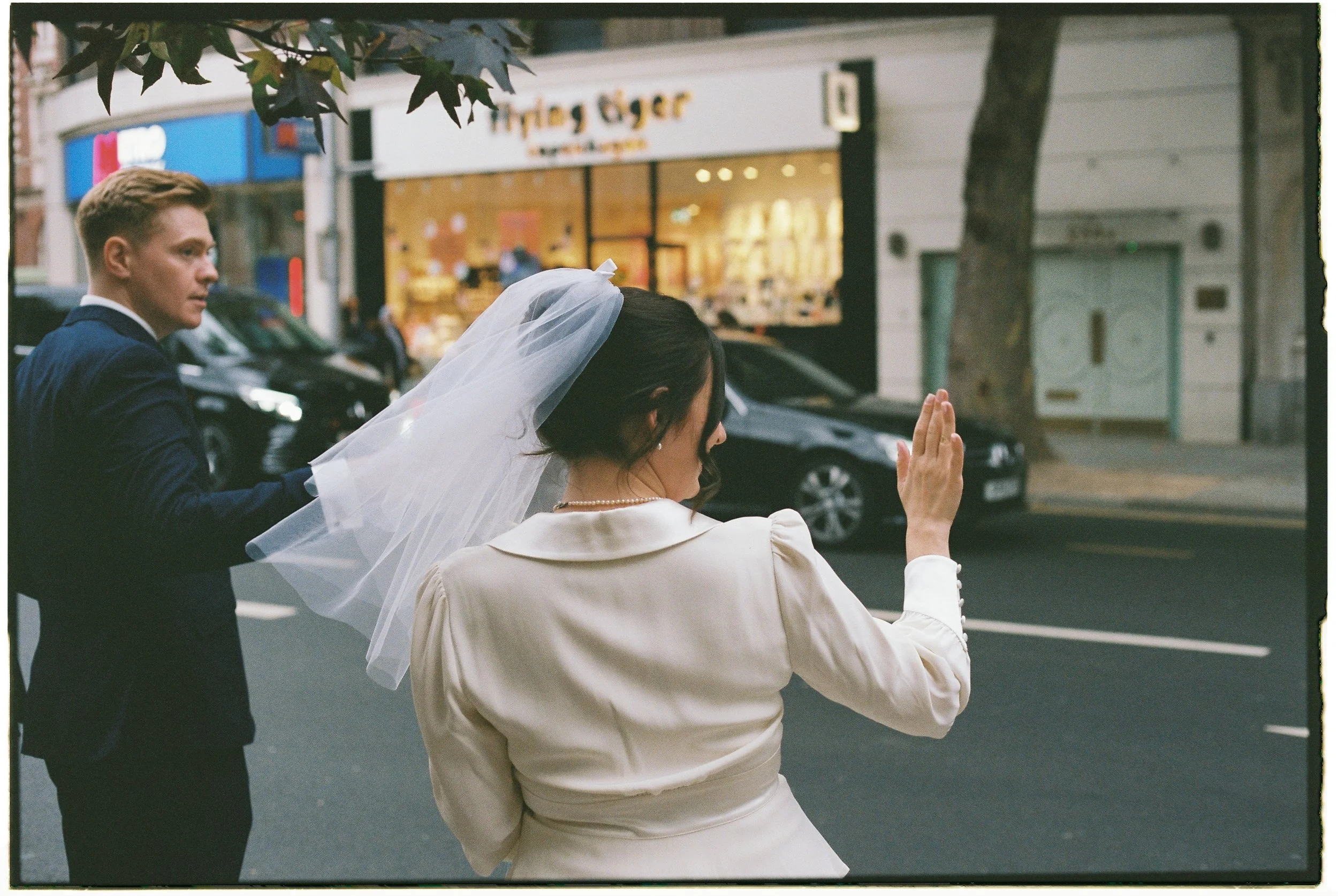 A bride waving her hand on a city street with a groom walking behind her, buildings, cars, and storefronts in the background.