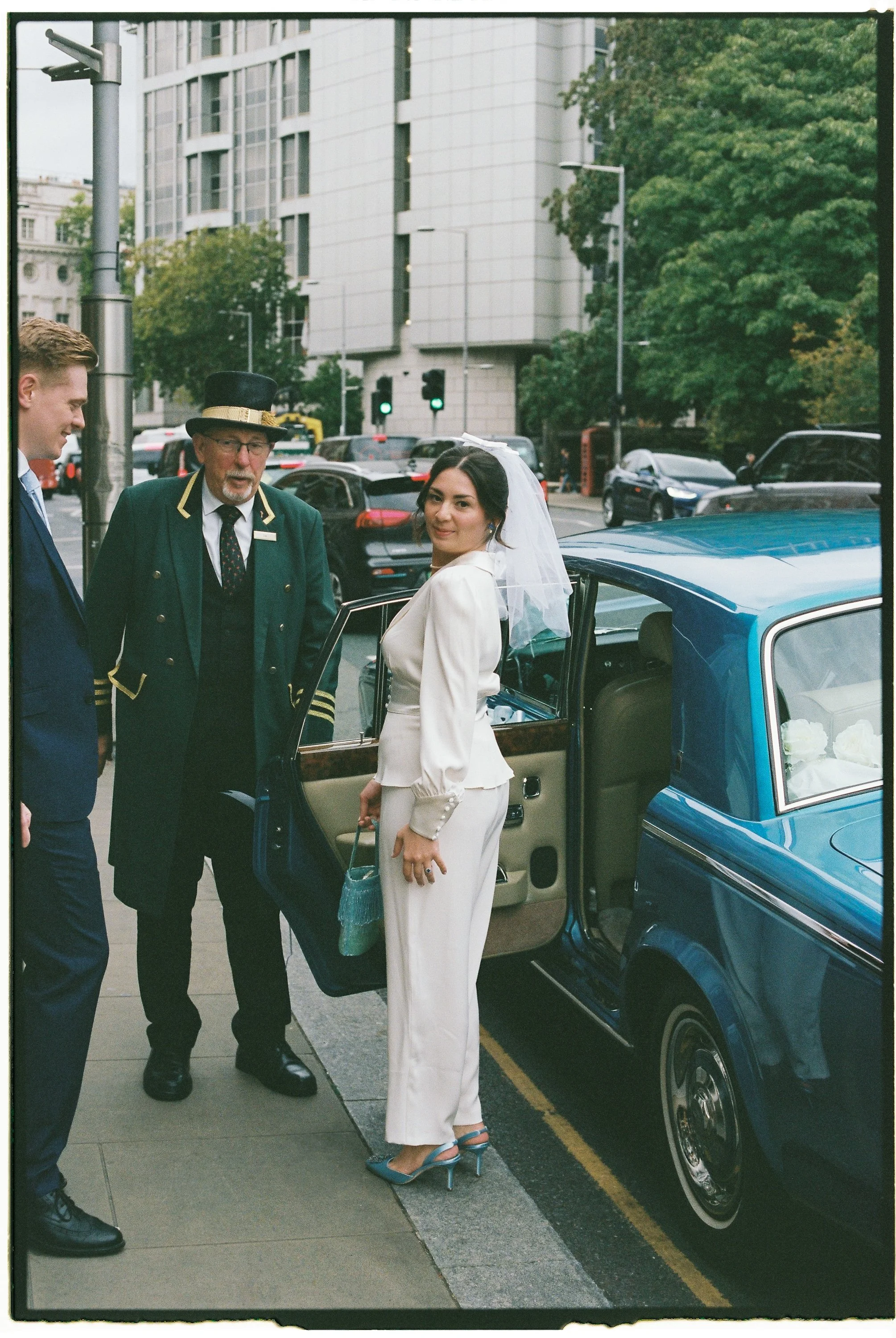 A woman in a white satin suit with a veil stands by a classic blue car, smiling at the camera, while two men in suits and a man in a vintage uniform converse nearby on a city street with tall buildings and traffic lights.