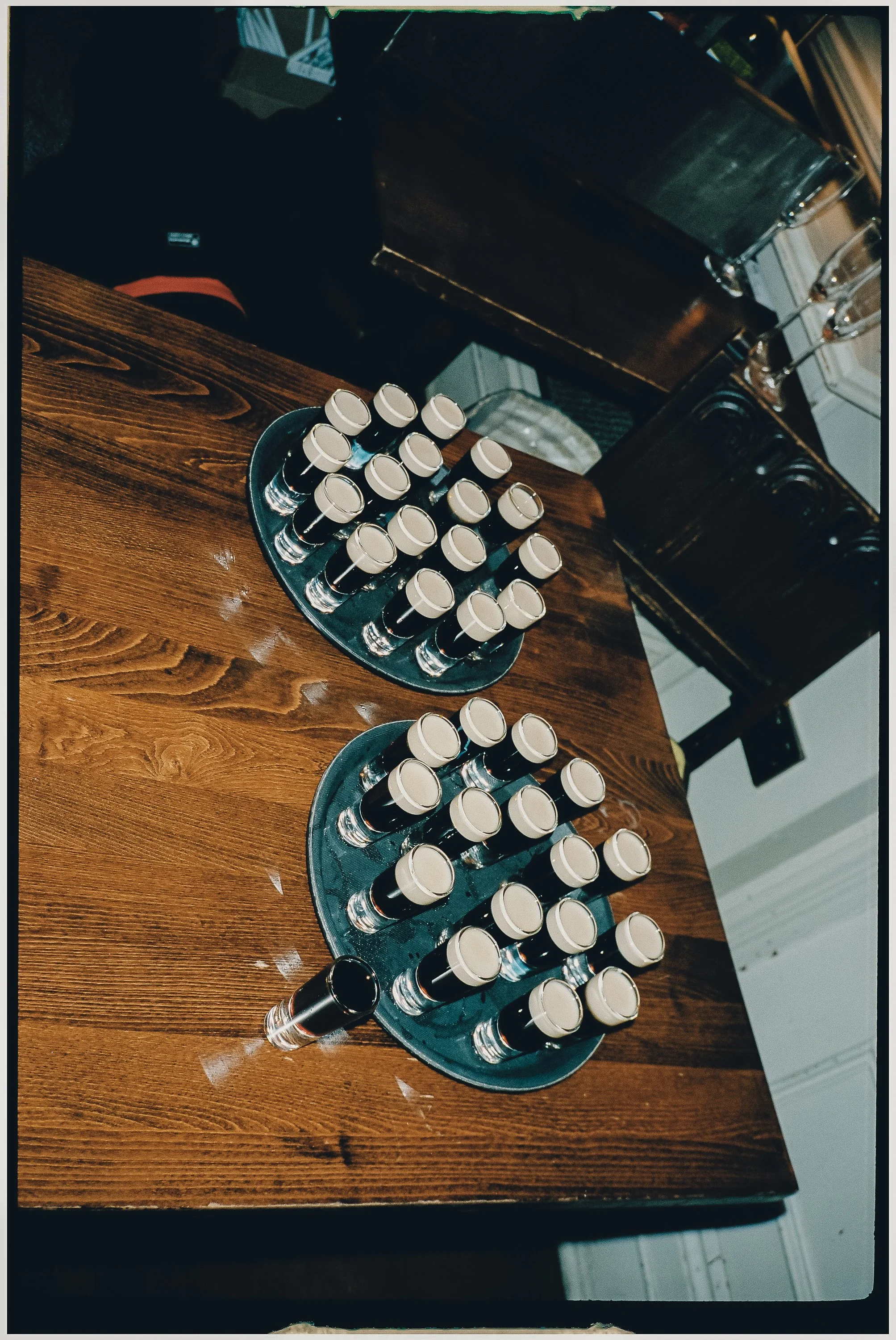 Two black trays each holding multiple small shot glasses filled with a dark beverage, placed on a wooden table.