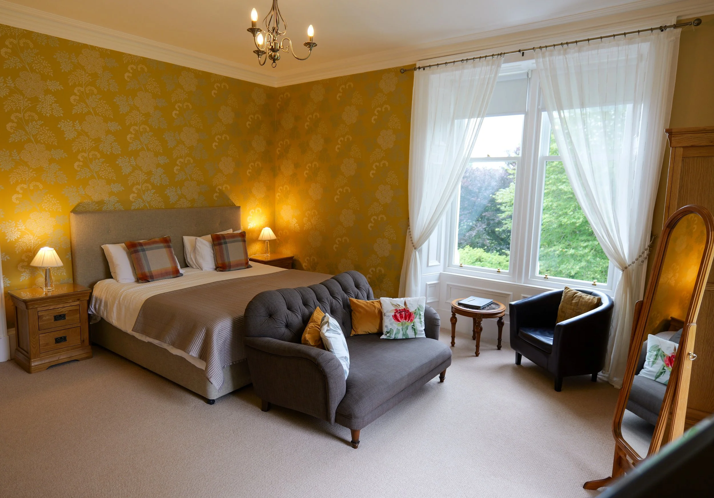 A cozy bedroom with a large bed, yellow floral wallpaper, and a window with white curtains showing a lush green view outside. There are nightstands with lamps, a small dark sofa with pillows, a round side table, and a full-length mirror.