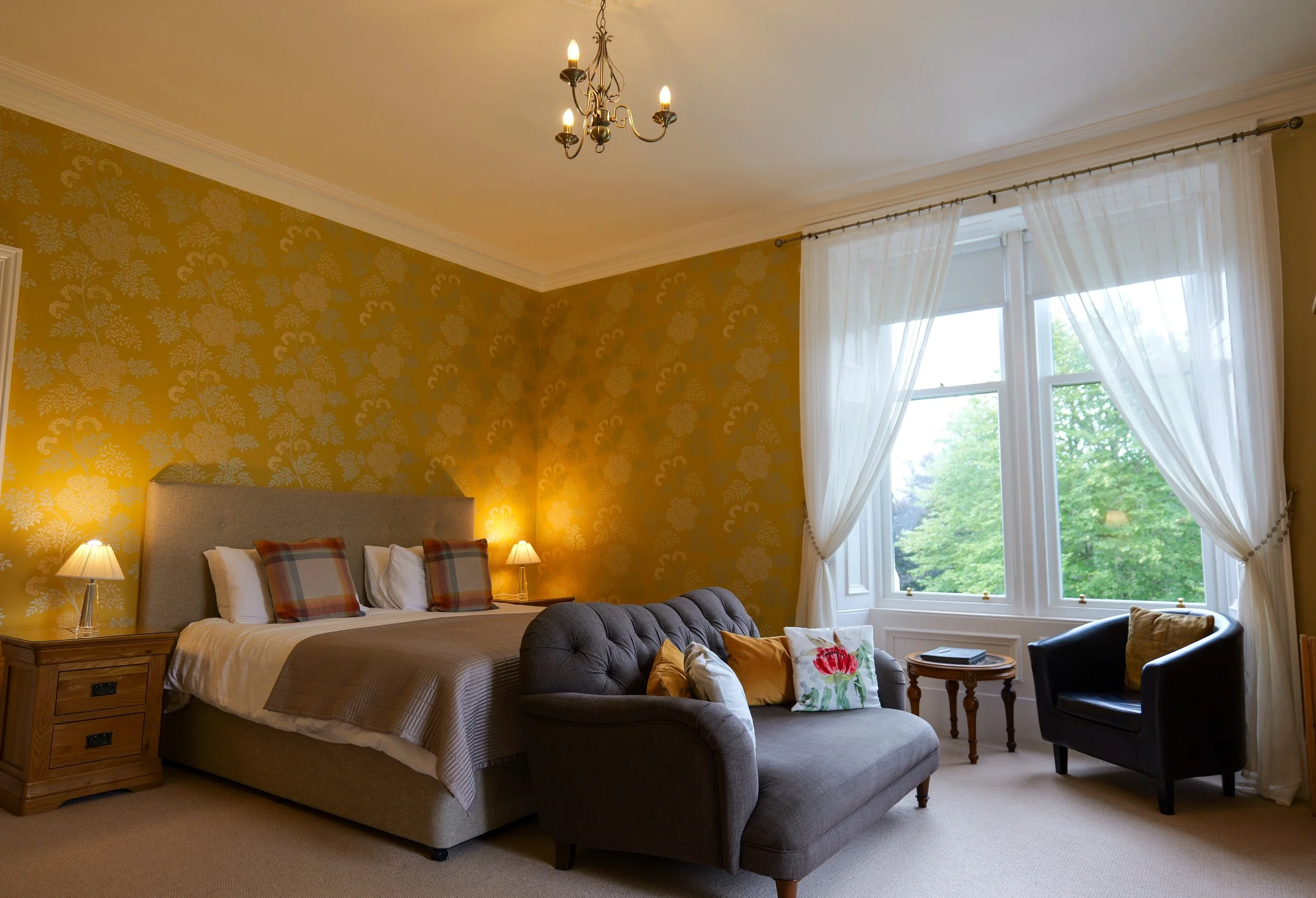 A cozy bedroom with a beige upholstered bed, plaid pillows, nightstands with lamps, a gray tufted loveseat with pillows, a round wooden side table, a black armchair, and large windows with white curtains overlooking green trees.