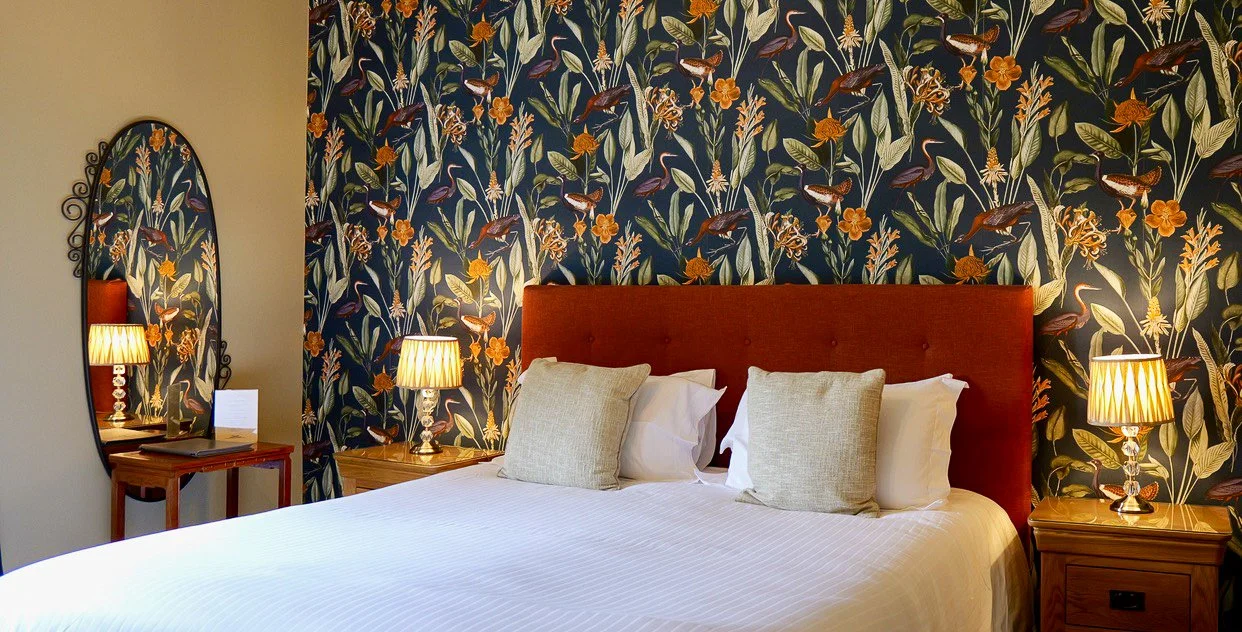 A hotel bedroom with a large bed featuring beige pillows and a red headboard, flanked by two wooden nightstands with matching lamps, and a wall decorated with tropical-patterned wallpaper.
