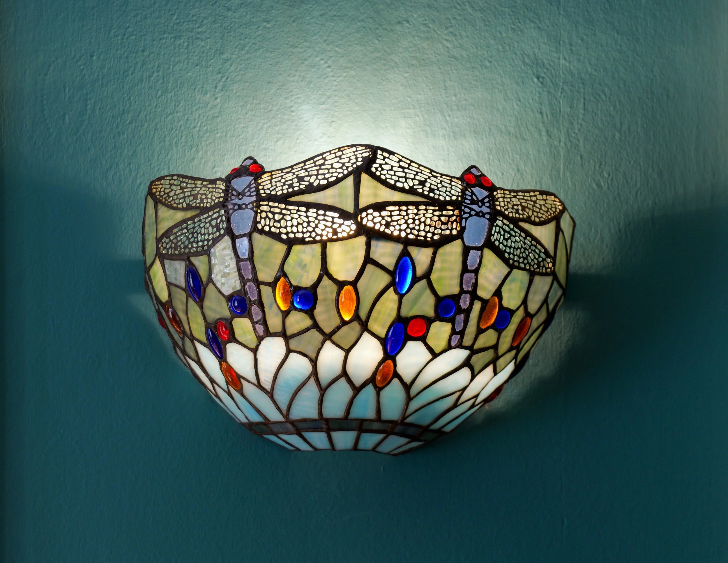 Tiffany-style wall sconce with dragonfly design, stained glass body, and colored glass accents, mounted on a teal wall.