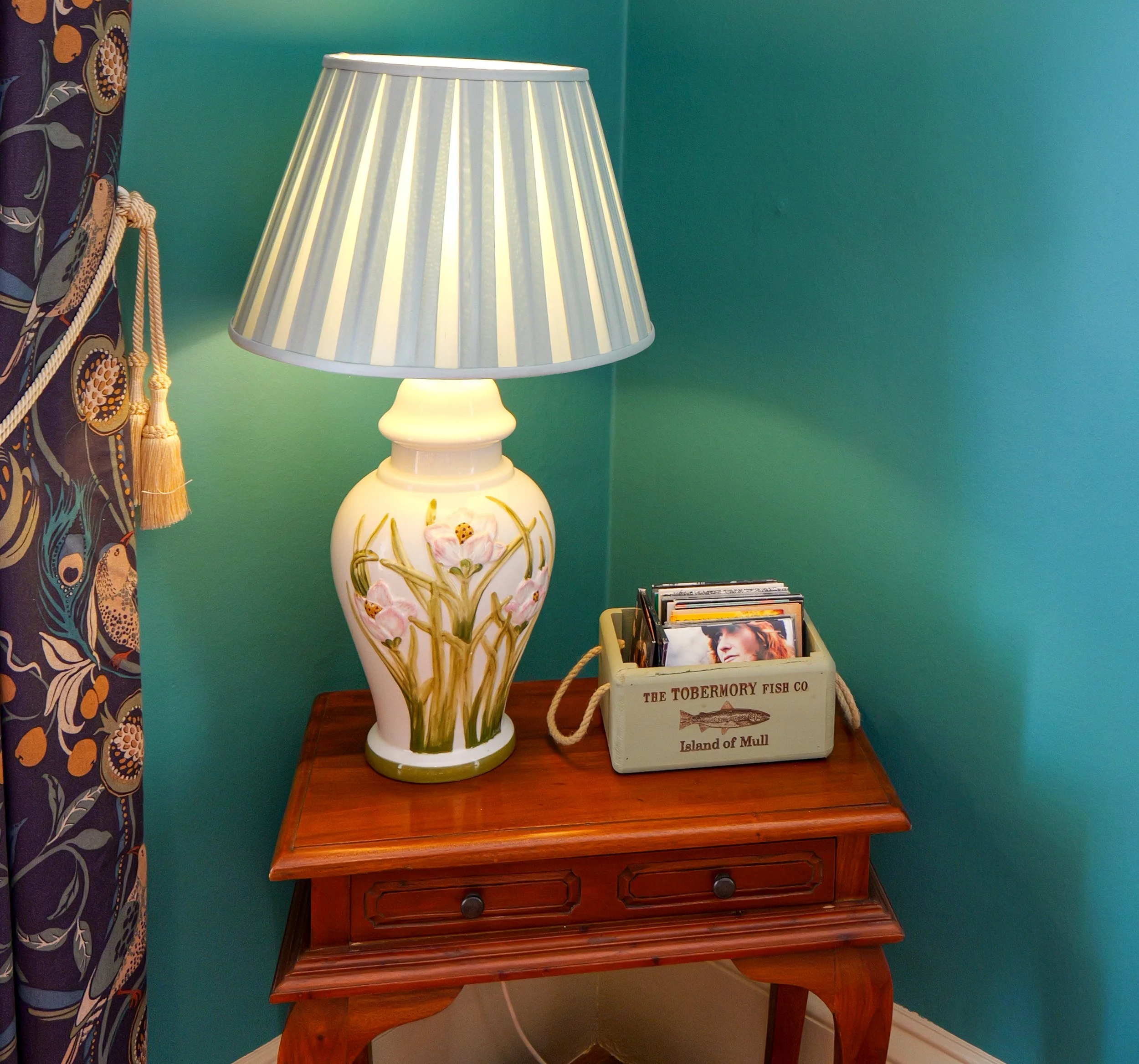 A wooden side table with a ceramic lamp and a white box filled with vinyl records in a cozy corner of a room.