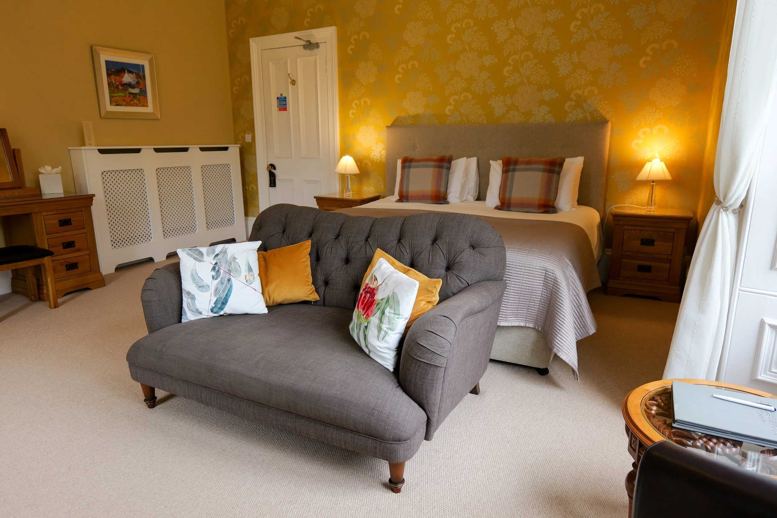 Hotel room with a bed, nightstands with lamps, a gray tufted loveseat with decorative pillows, and a small round table with a notepad and pen, featuring warm yellow wallpaper with floral patterns.